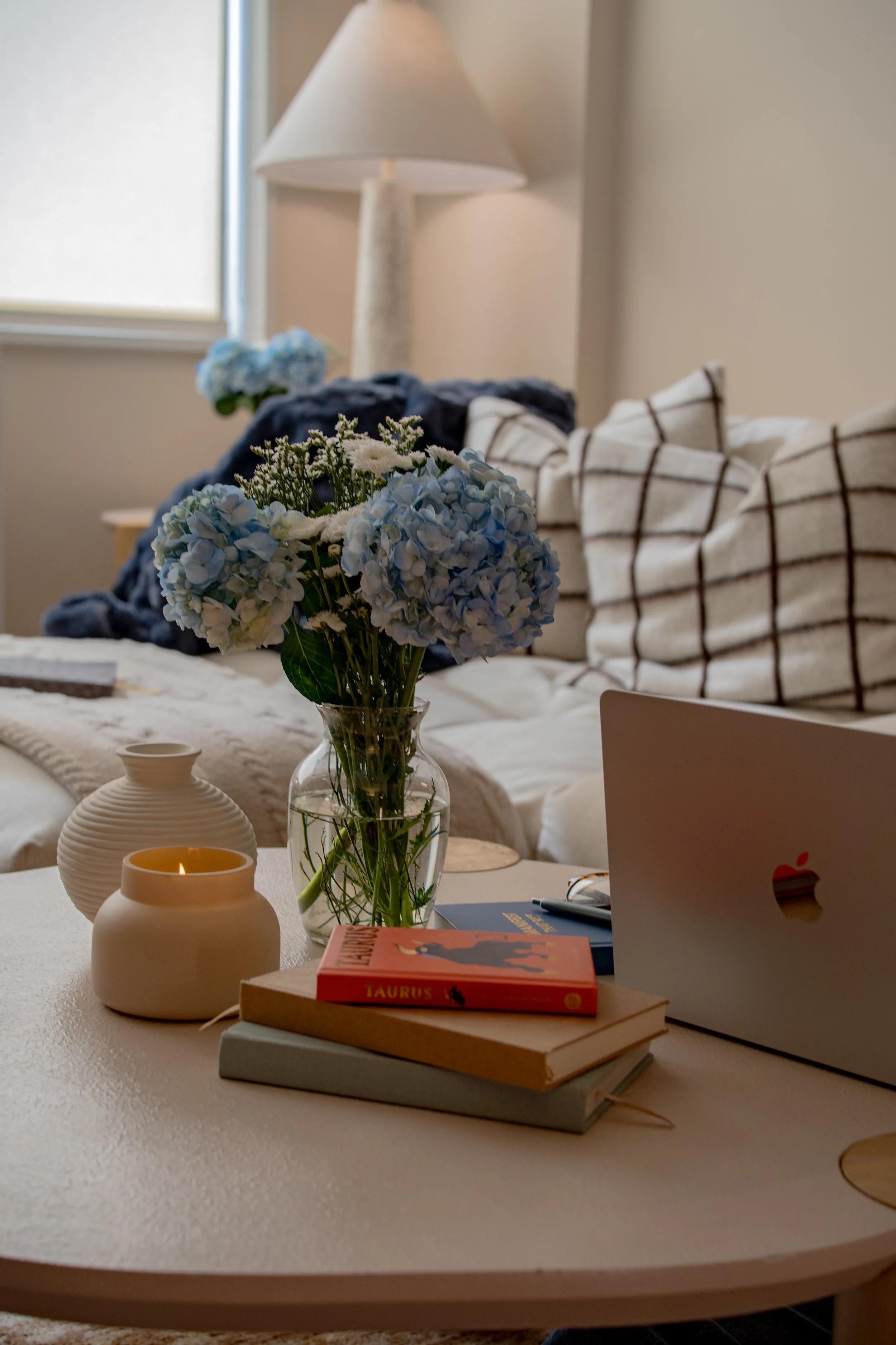 A bouquet of light blue hydrangeas in a glass vase is placed on a white table. On the table are two white vases, one with a candle inside, a stack of books with a red book titled "Taurus" on top, a pen, and an open silver laptop. In the background, t