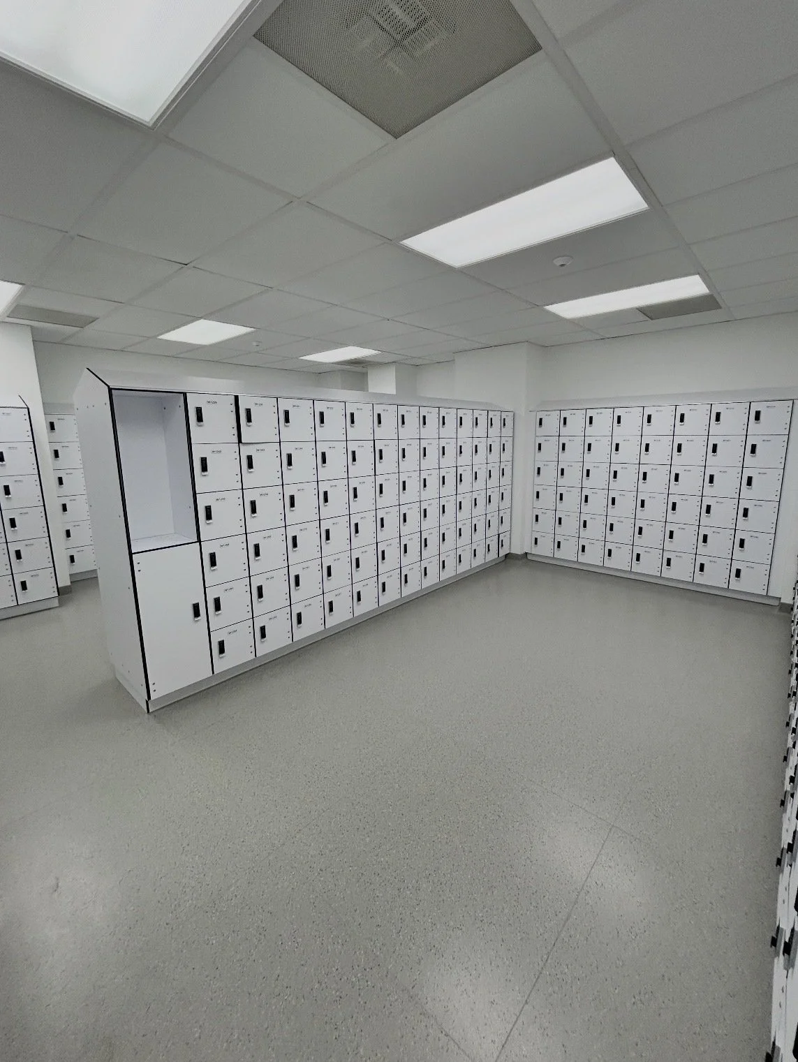 Room with multiple white lockers arranged along the walls, some lockers have locks. The ceiling has fluorescent lights and an air vent.