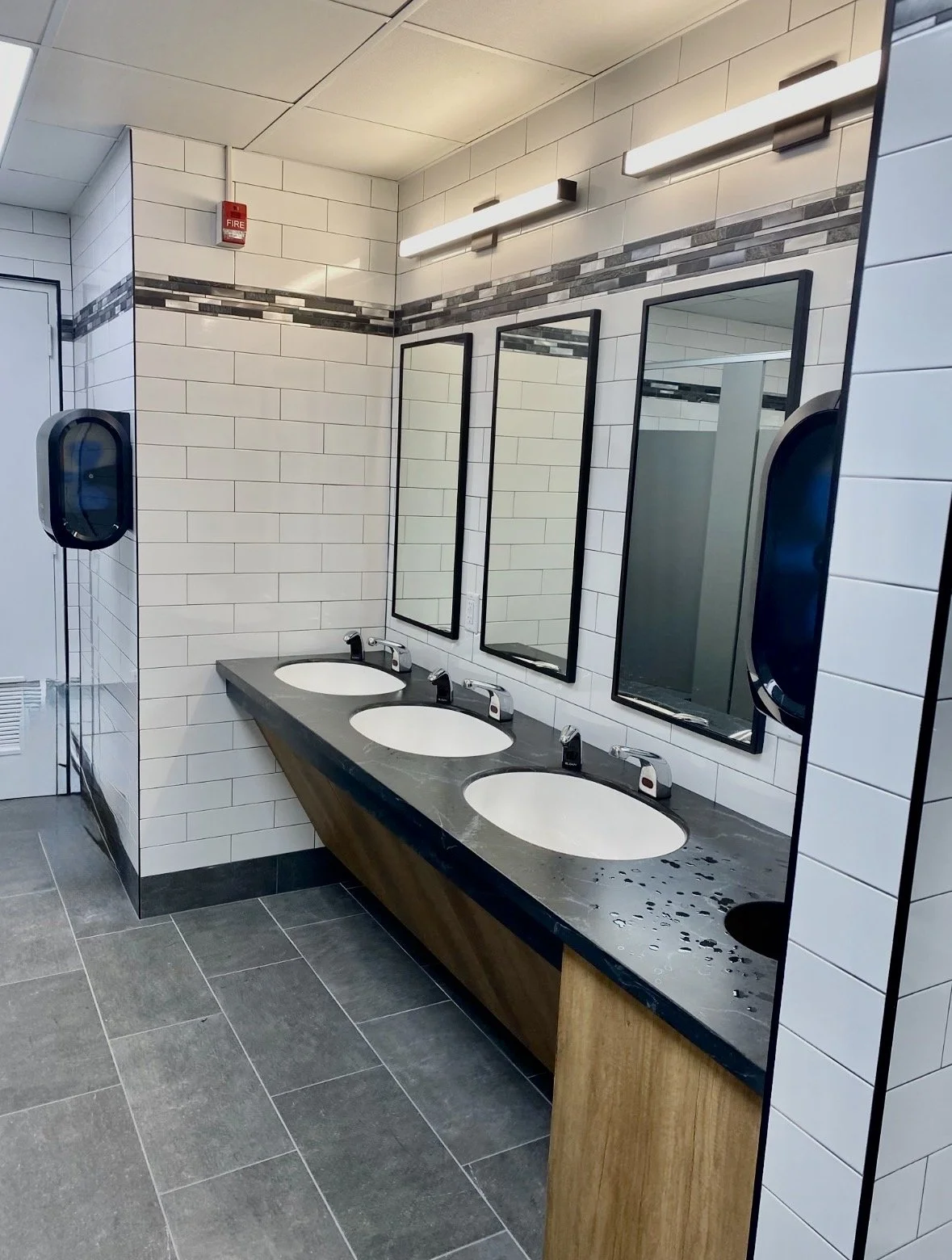 Public men's restroom with three sinks, mirrors, soap dispensers, and a paper towel or hand dryer on each side, tiled walls with decorative border, and gray tiled floor.
