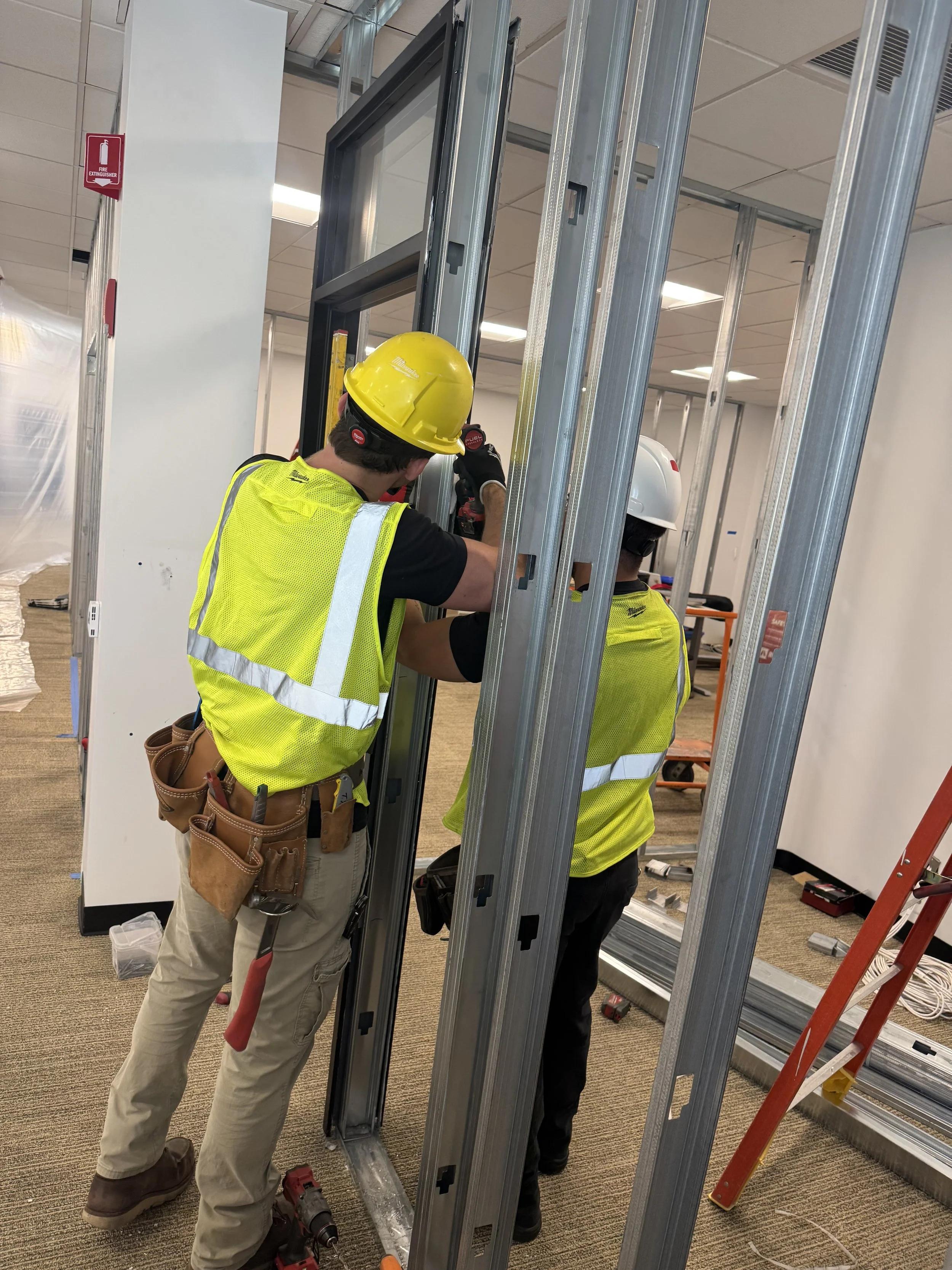 Two construction workers installing metal framing for interior walls in an office space. One worker is wearing a yellow hard hat and is working with a drill, while the other worker is wearing a white hard hat and appears to be assisting. They are surrounded by metal studs and tools, with a ladder nearby.