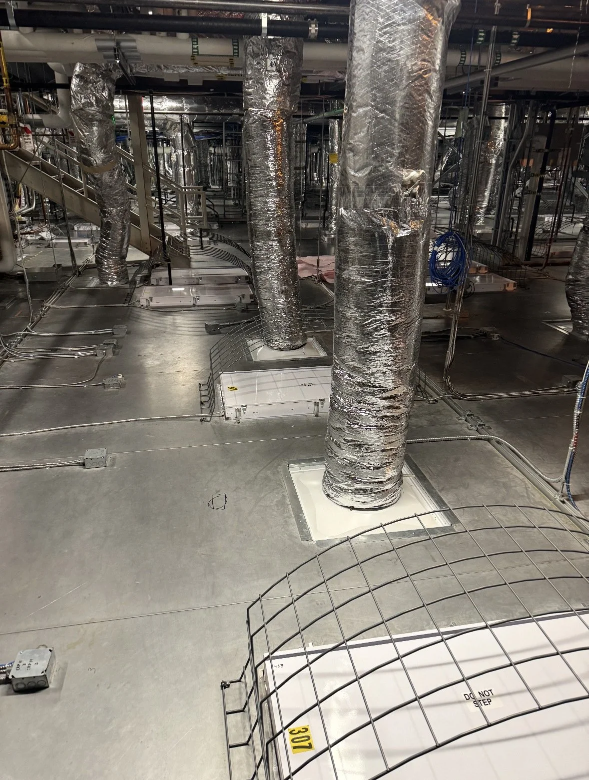 View of an industrial mechanical room showing large insulated ducts, electrical conduits, and various equipment underneath a metal floor structure.