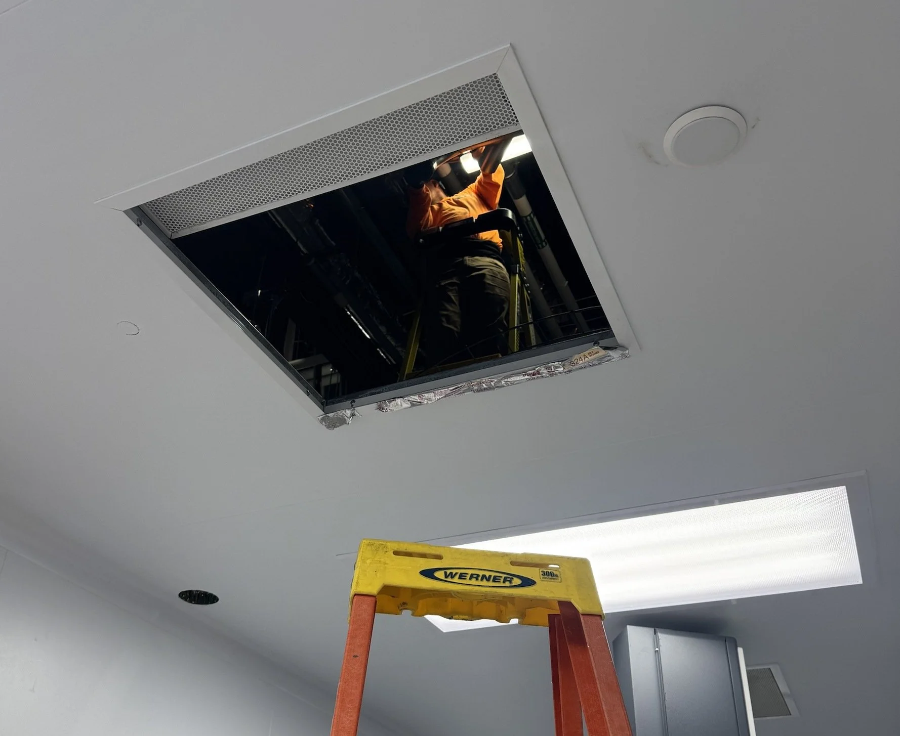 A worker in an orange shirt and dark pants standing on a yellow Werner ladder, installing or repairing a ceiling vent or air conditioning unit in a drop ceiling in an office or commercial building. The ceiling tiles have been removed around the vent.