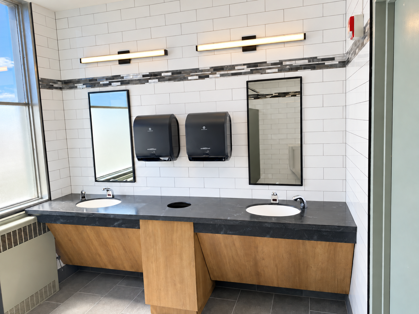 Restroom with a double sink vanity, black countertop, two rectangular mirrors, two black paper towel dispensers, white tiled walls with black and gray decorative tiles, and a large window.