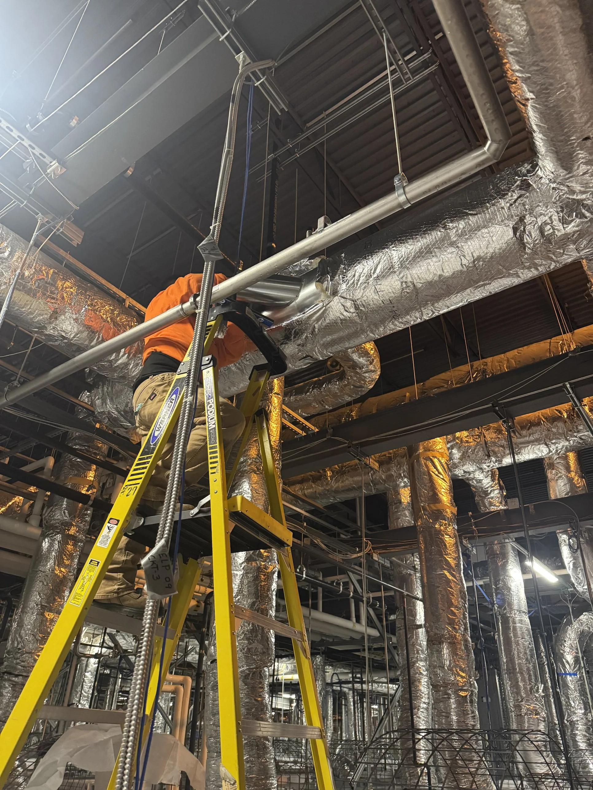 A construction worker is standing on a yellow ladder, working on the ceiling of an industrial or commercial building. The ceiling has numerous metallic ducts and piping, some wrapped in insulation, with various support structures and wires visible.