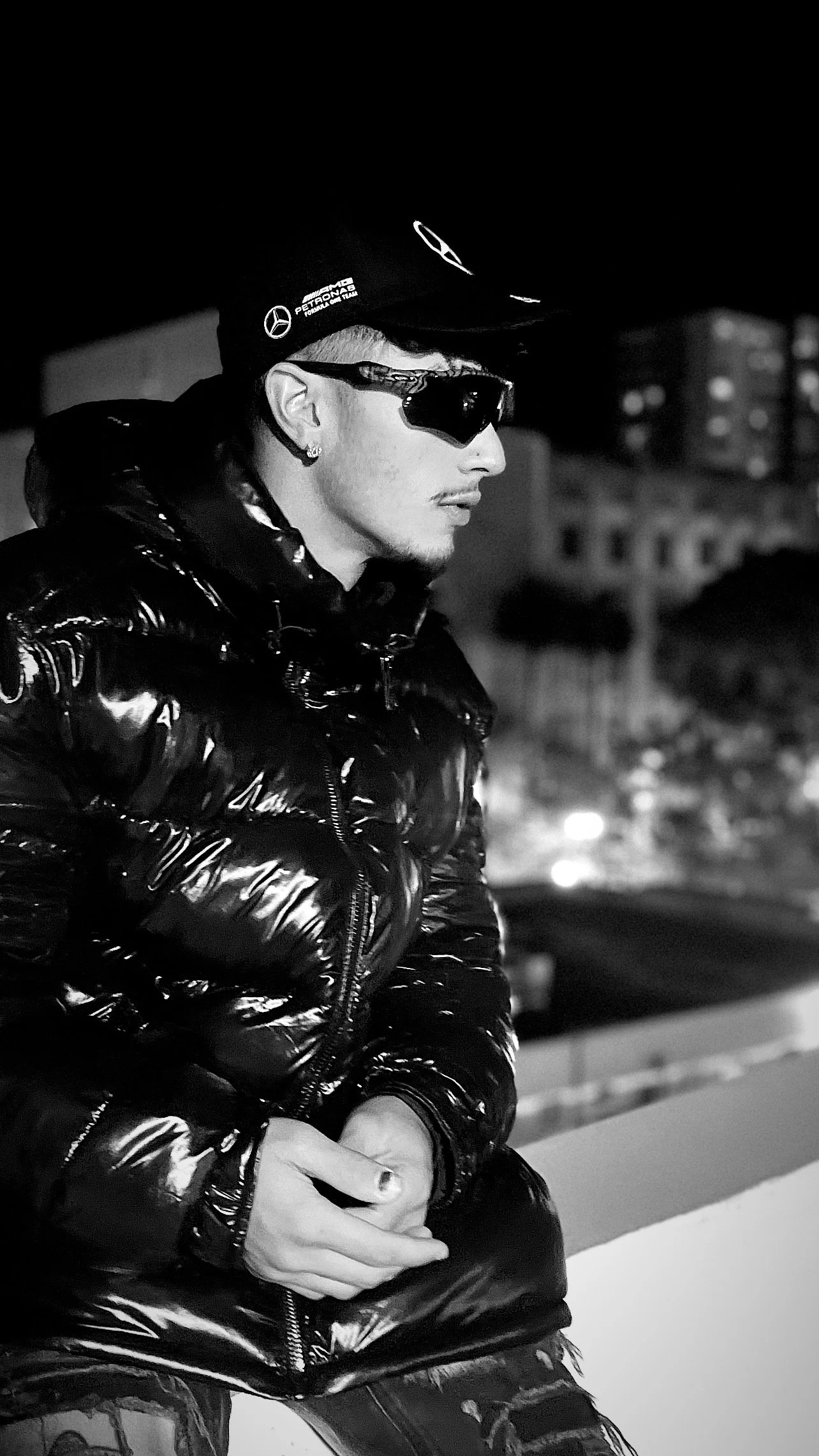A man with tattoos on his hands, wearing sunglasses, a beanie, and a shiny puffer jacket, sitting outdoors at night.