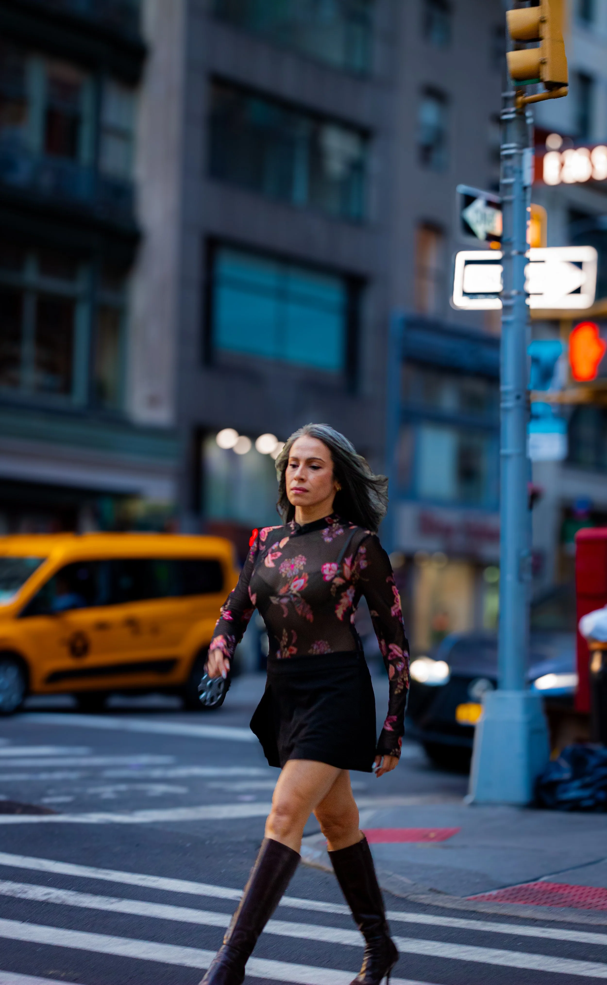 A woman with dark hair wearing a sheer floral top, black shorts, and tall black boots walking across a city crosswalk during dusk. Yellow taxis and city buildings are visible in the background.