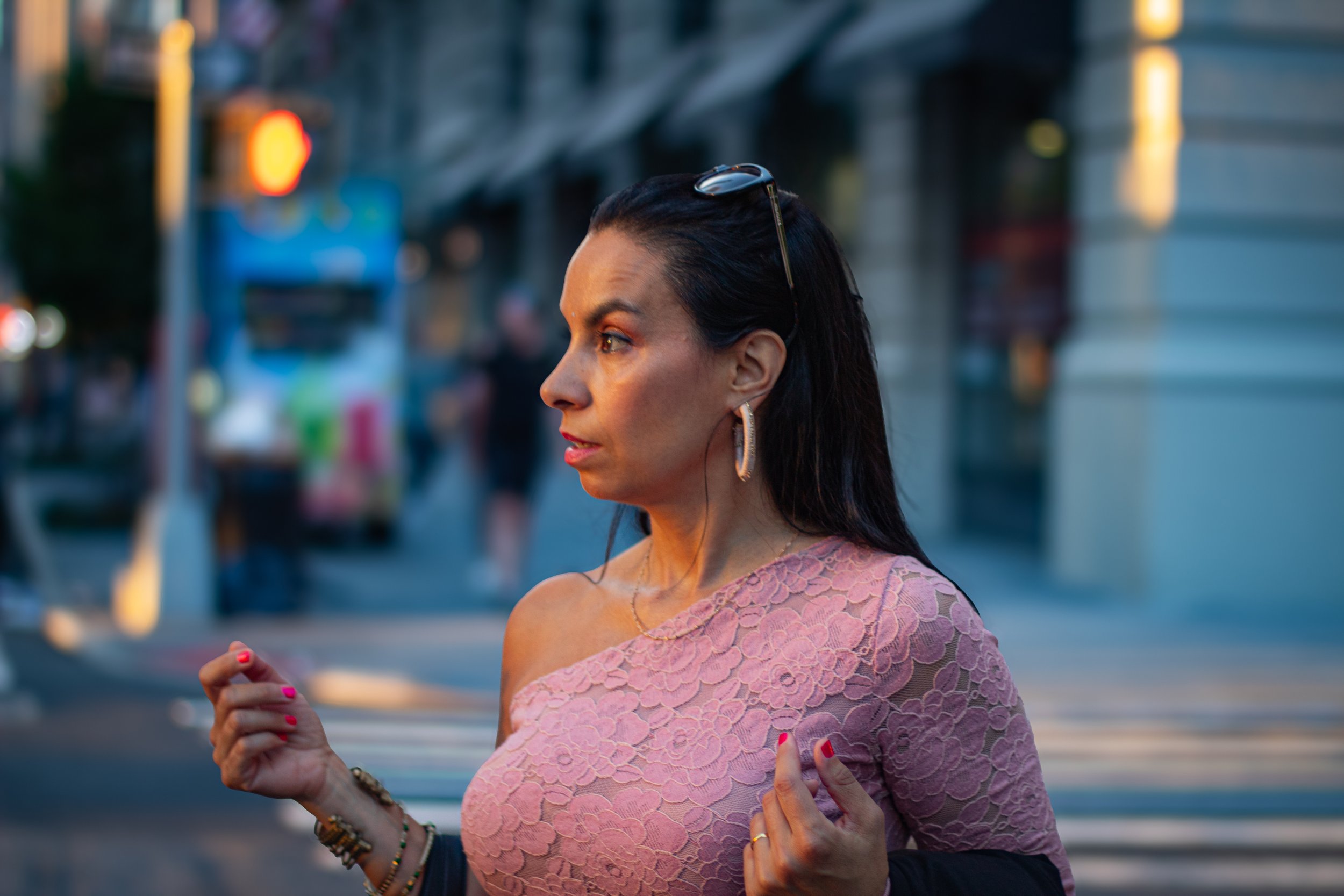 A woman with dark hair in a bun, wearing a pink lace top, earrings, and multiple bracelets, is walking outdoors among blurred cityscape background, captured in profile.