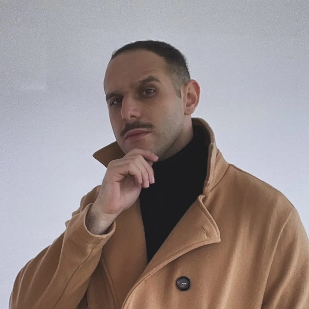 A man with short dark hair, mustache, wearing a black turtleneck and tan coat, posing against a plain, light-colored background.