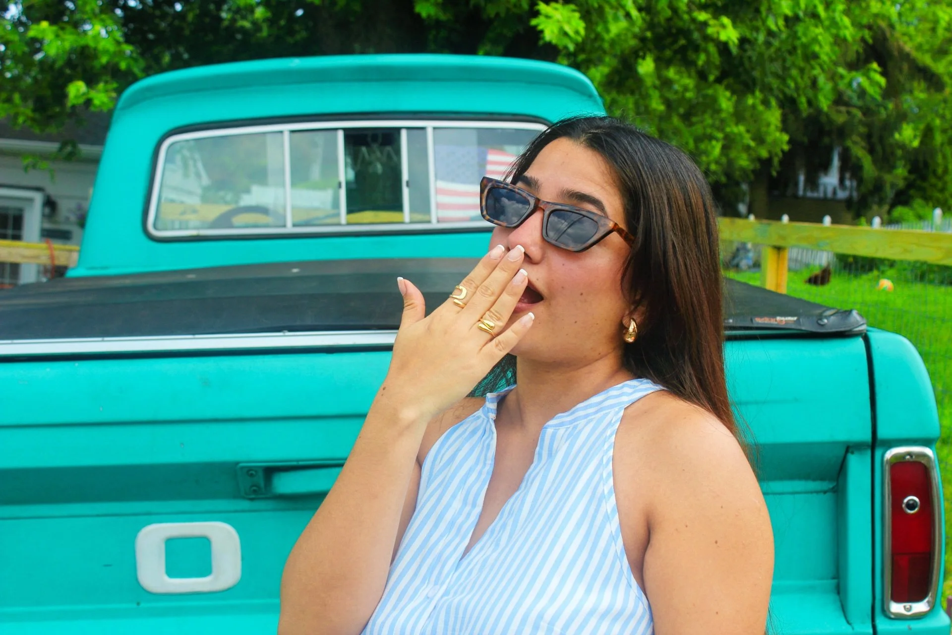 A woman with dark hair wearing sunglasses and gold jewelry poses in front of a vintage turquoise pickup truck. The woman is wearing a blue and white striped sleeveless top and has her hand near her face, with her nails painted white. The background shows green trees and part of a house with a white fence.