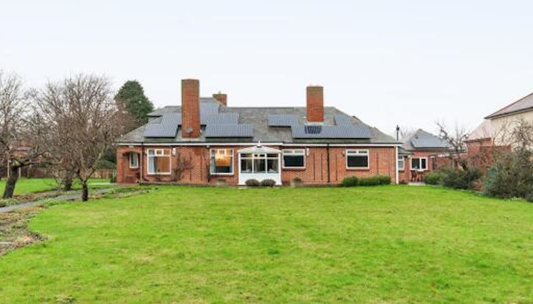 A large brick house with a dark roof and multiple chimneys, surrounded by a lush green lawn and sparse trees.