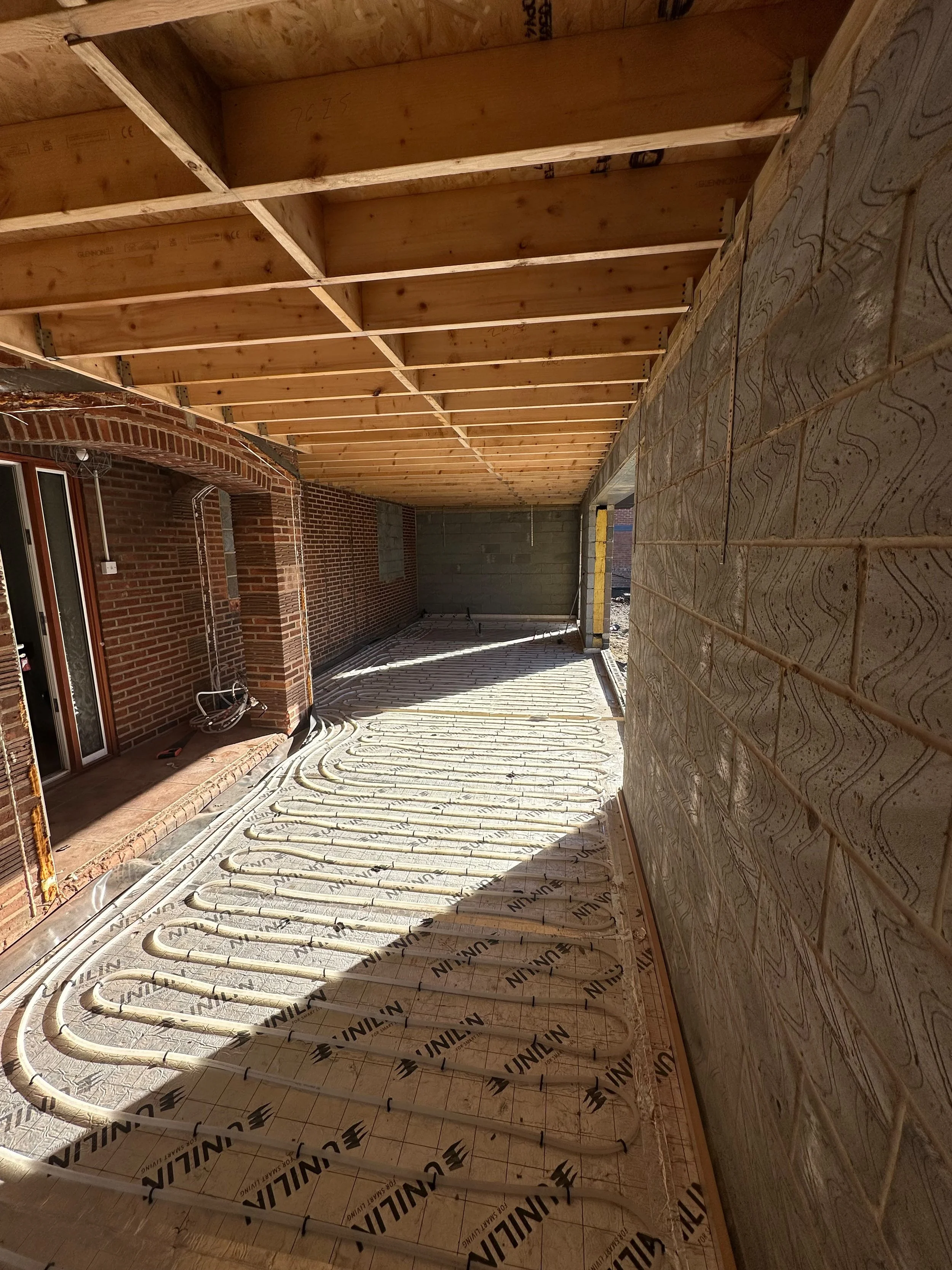 Construction site with a wooden ceiling framework and heated flooring hydronic tubing installed on the floor, next to a brick wall with an arched brick doorway.
