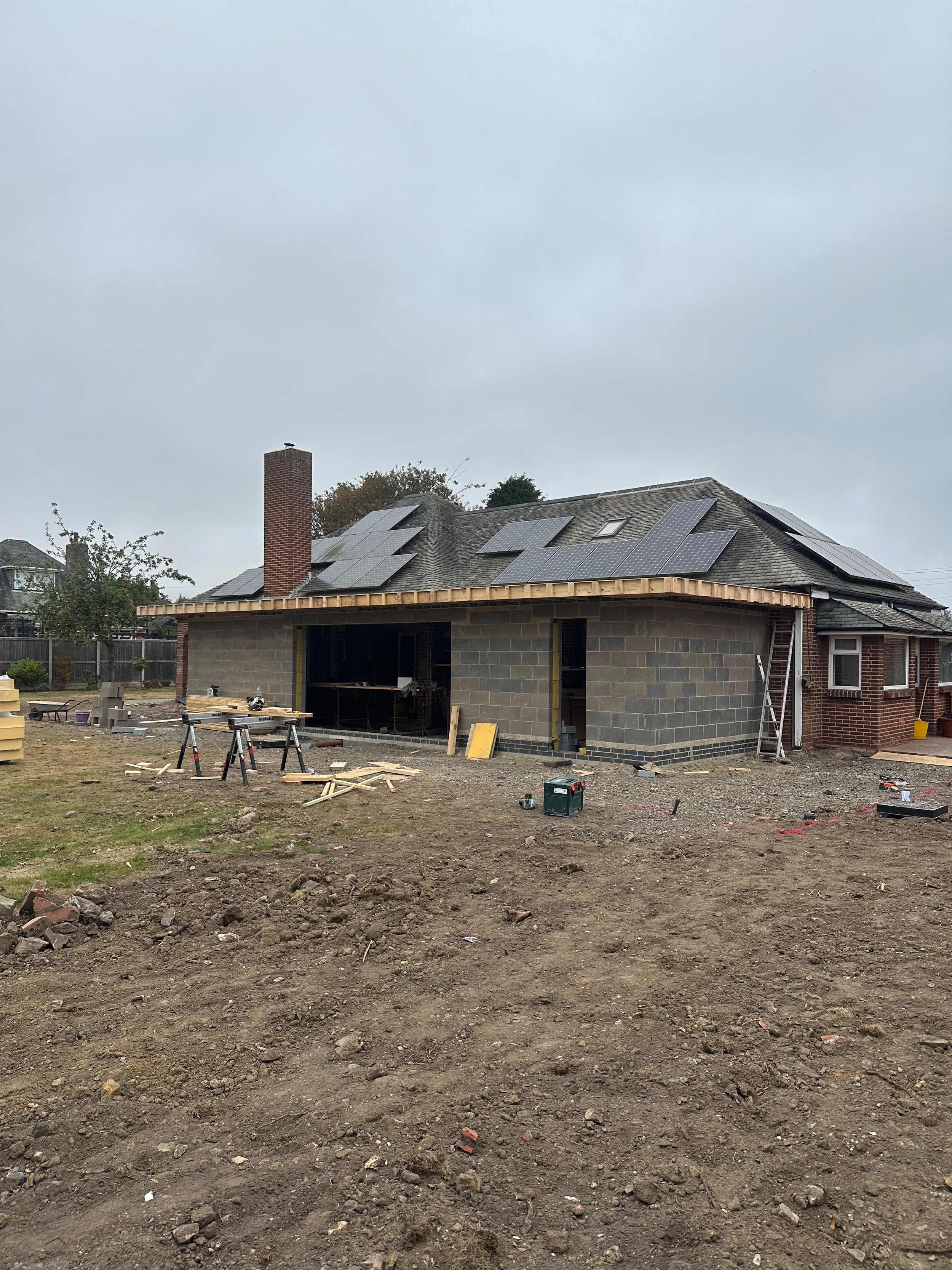 A house under construction with solar panels on the roof, surrounded by construction tools and materials on the ground, with a cloudy sky overhead.