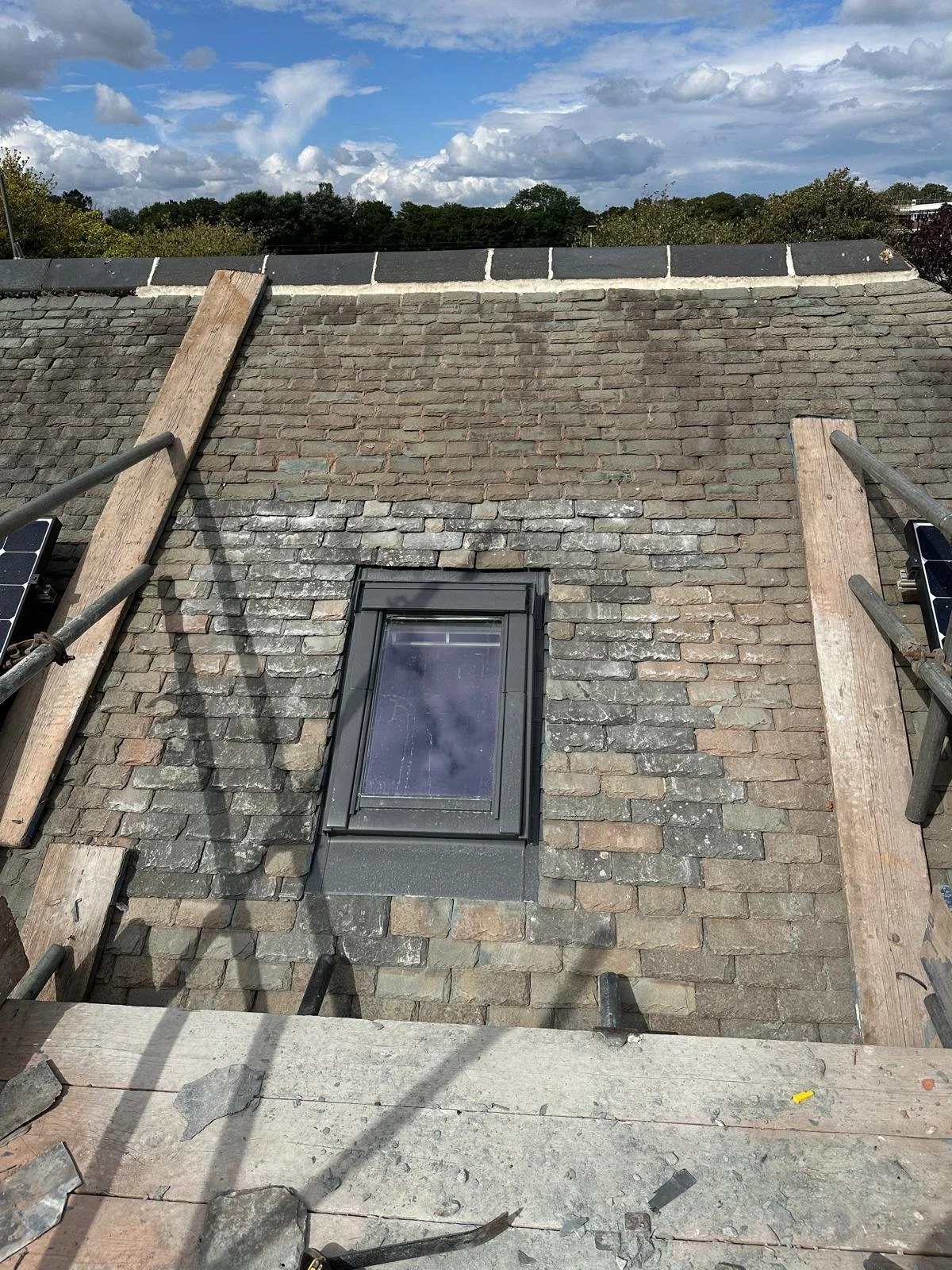 Roof under repair with scaffolding, wooden planks, and a skylight window. Cloudy sky and trees in the background.
