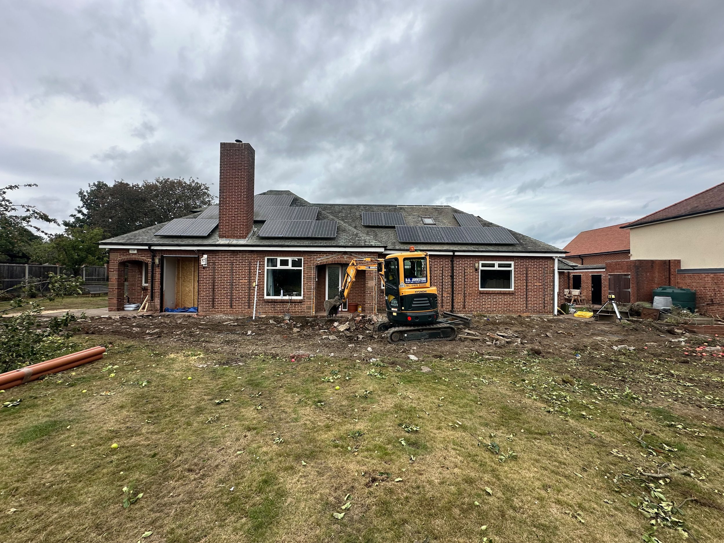 A house with solar panels on the roof under a cloudy sky, currently undergoing renovation, with construction equipment and debris in the yard.