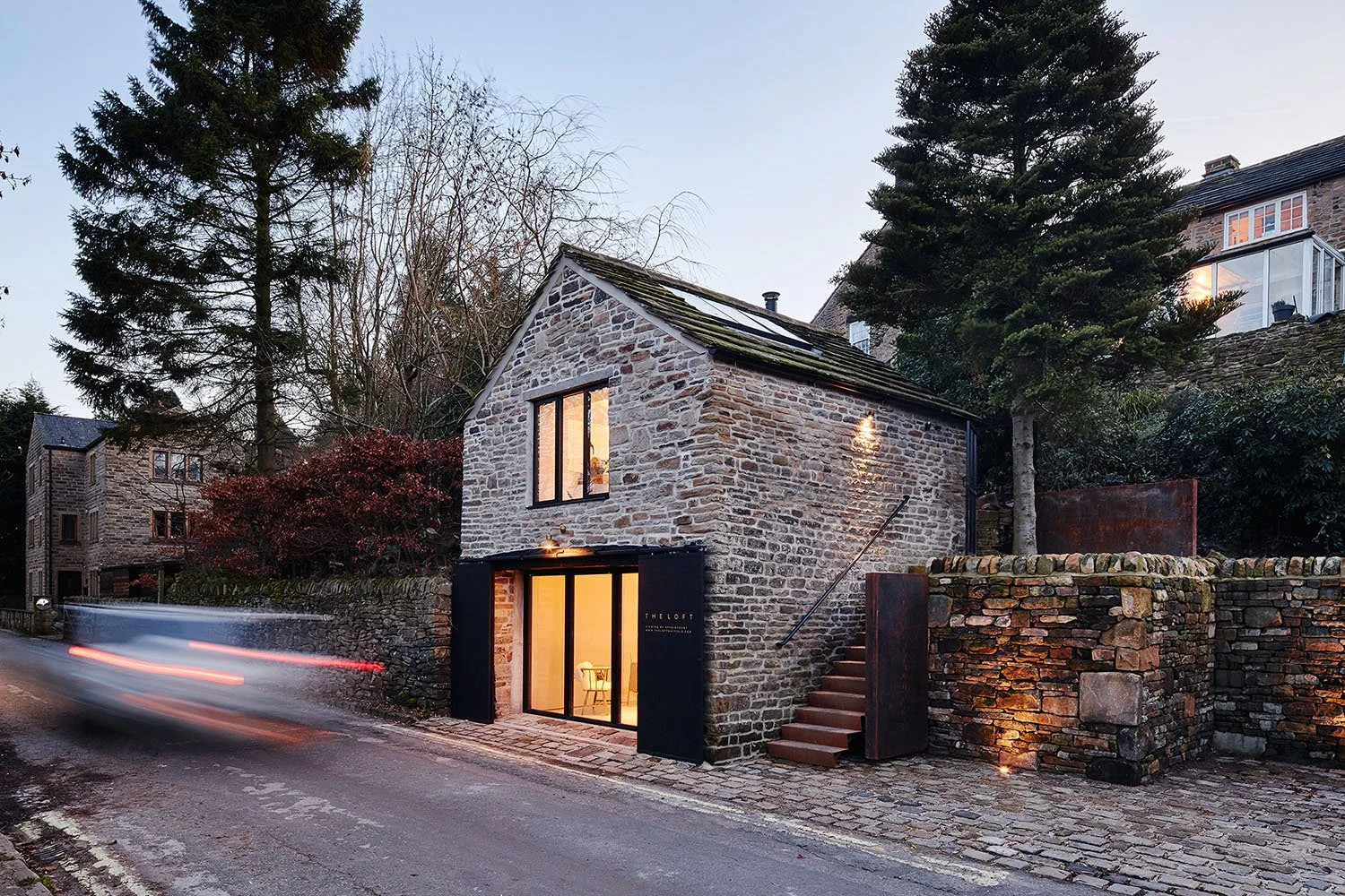 A stone house with large windows on a cobblestone street, with trees and other stone buildings nearby. The house has a modern look with black accents and warm lighting visible inside.