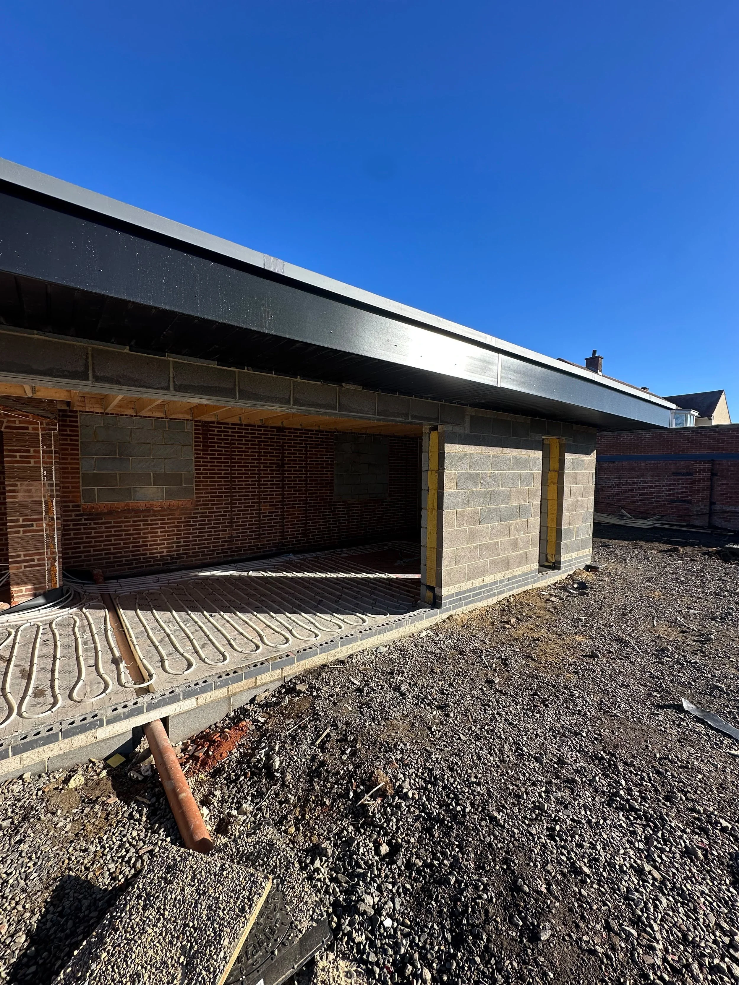 Construction site with incomplete brick walls and exposed concrete block columns, with underfloor heating pipes visible, and a clear blue sky above.