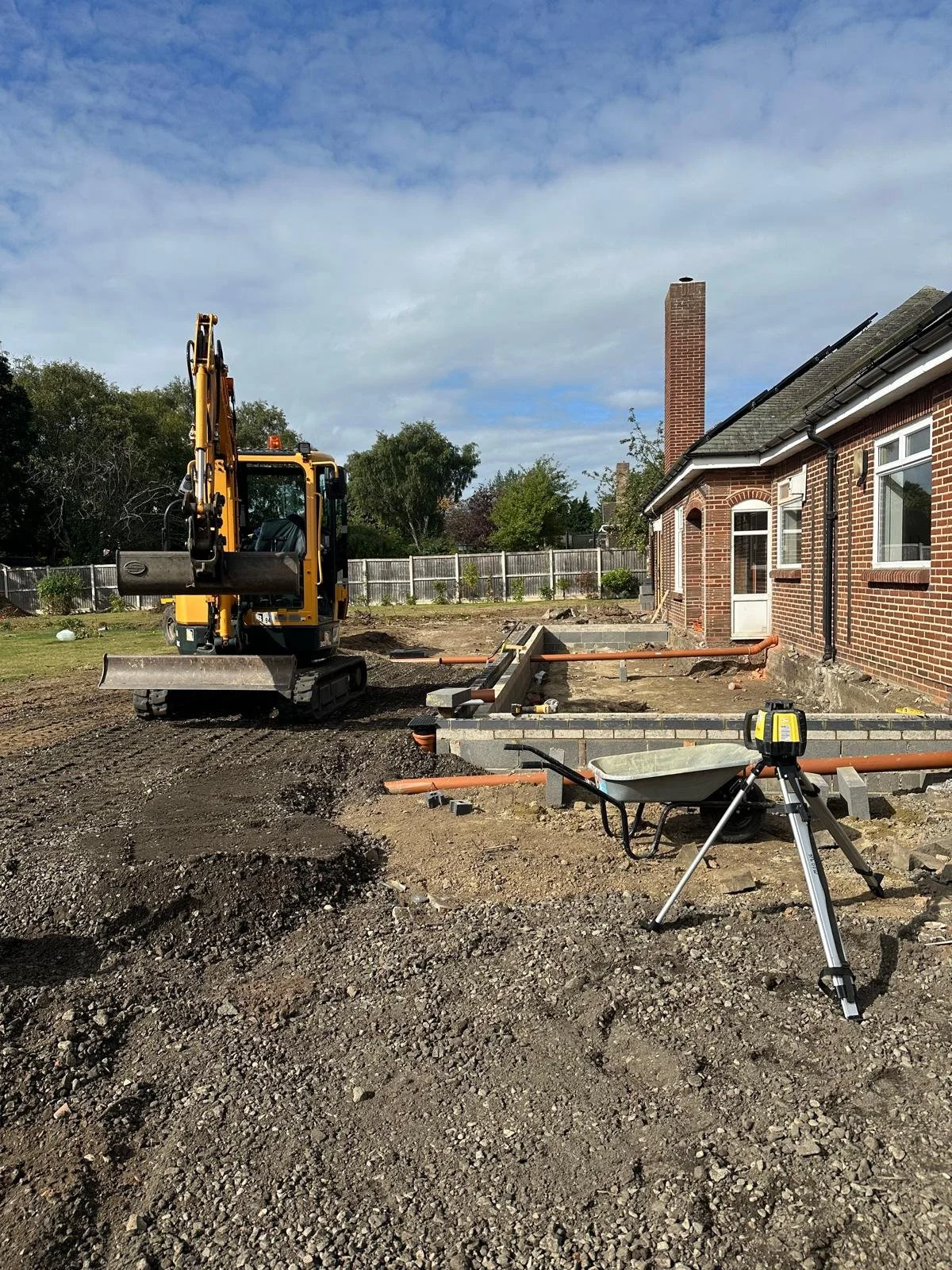 Construction site with a mini excavator, wheelbarrow, and construction tools in front of a brick house with a chimney, and a fenced backyard under a cloudy sky.