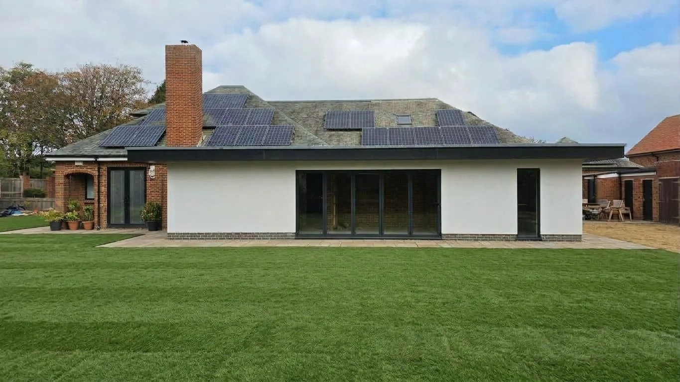 A modern house with solar panels on the roof, a brick chimney, a white exterior wall, large glass sliding doors, and a green lawn.