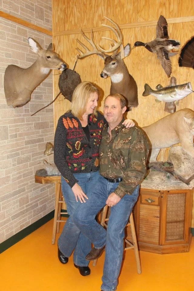 A man and woman smiling and embracing in a room with mounted deer and animal heads on the wooden wall behind them.