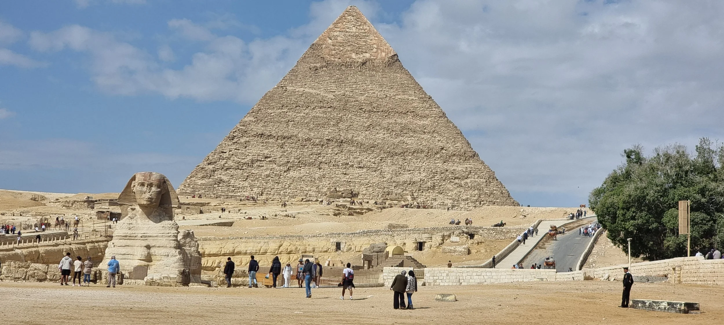 The Great Pyramid of Giza with the Sphinx in front, surrounded by tourists and a clear sky.