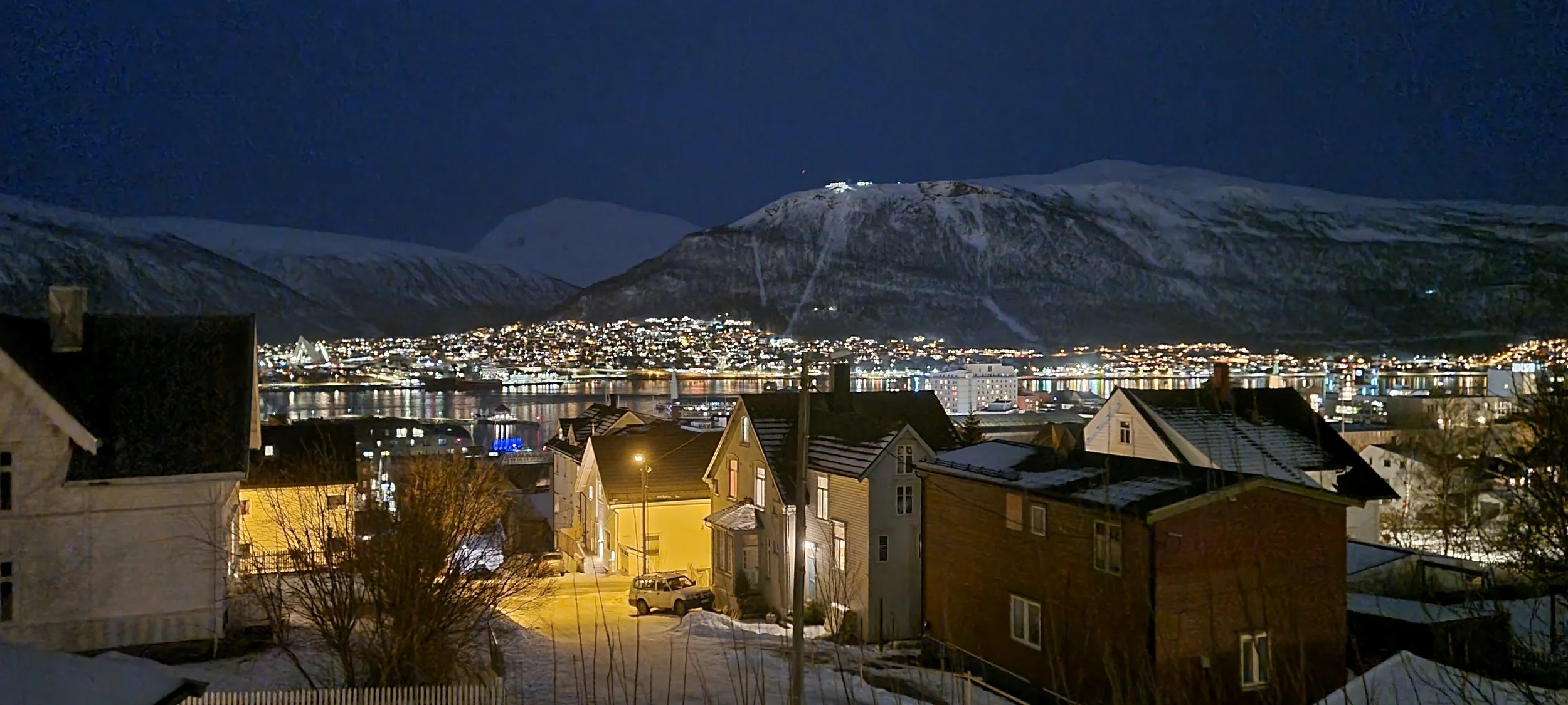 Nighttime view of a small town beside a body of water, with snow-covered mountains in the background. The town is lit up with streetlights and building lights, reflecting on the water. In the foreground, houses and cars are visible on snow-covered streets.