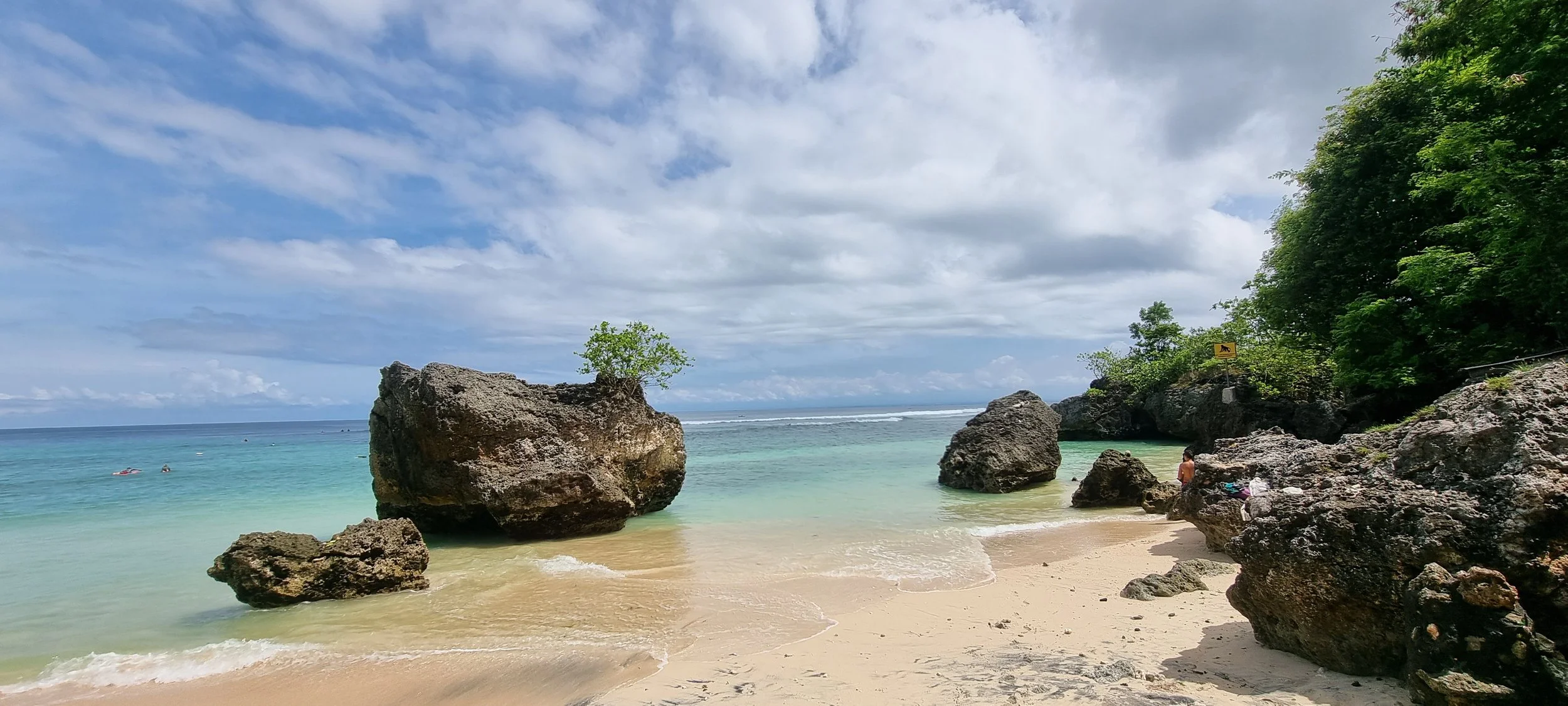 Tropical beach with large rocks, sandy shore, turquoise water, green trees, and cloudy sky.