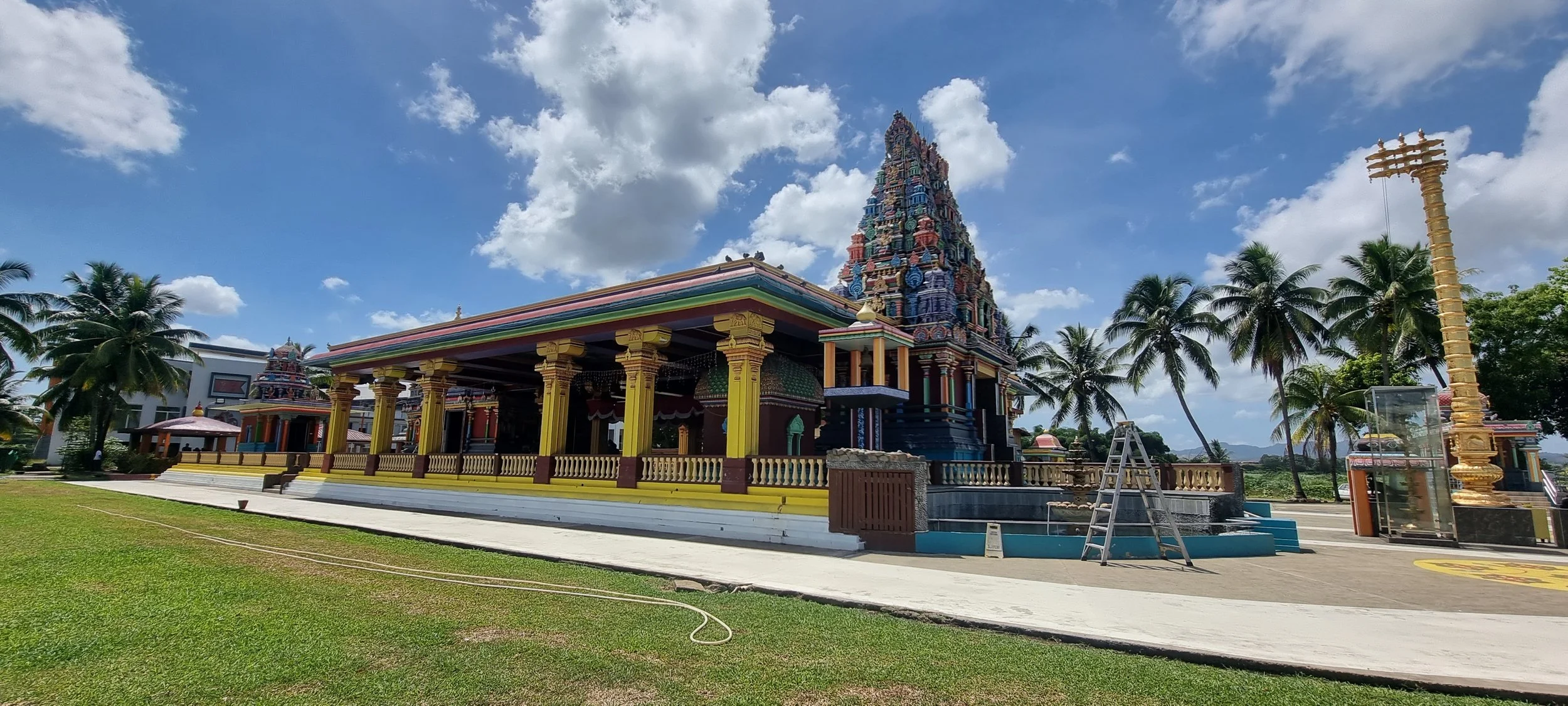 Colorful Hindu temple with ornate towers, surrounded by palm trees and a grassy yard under a partly cloudy sky.