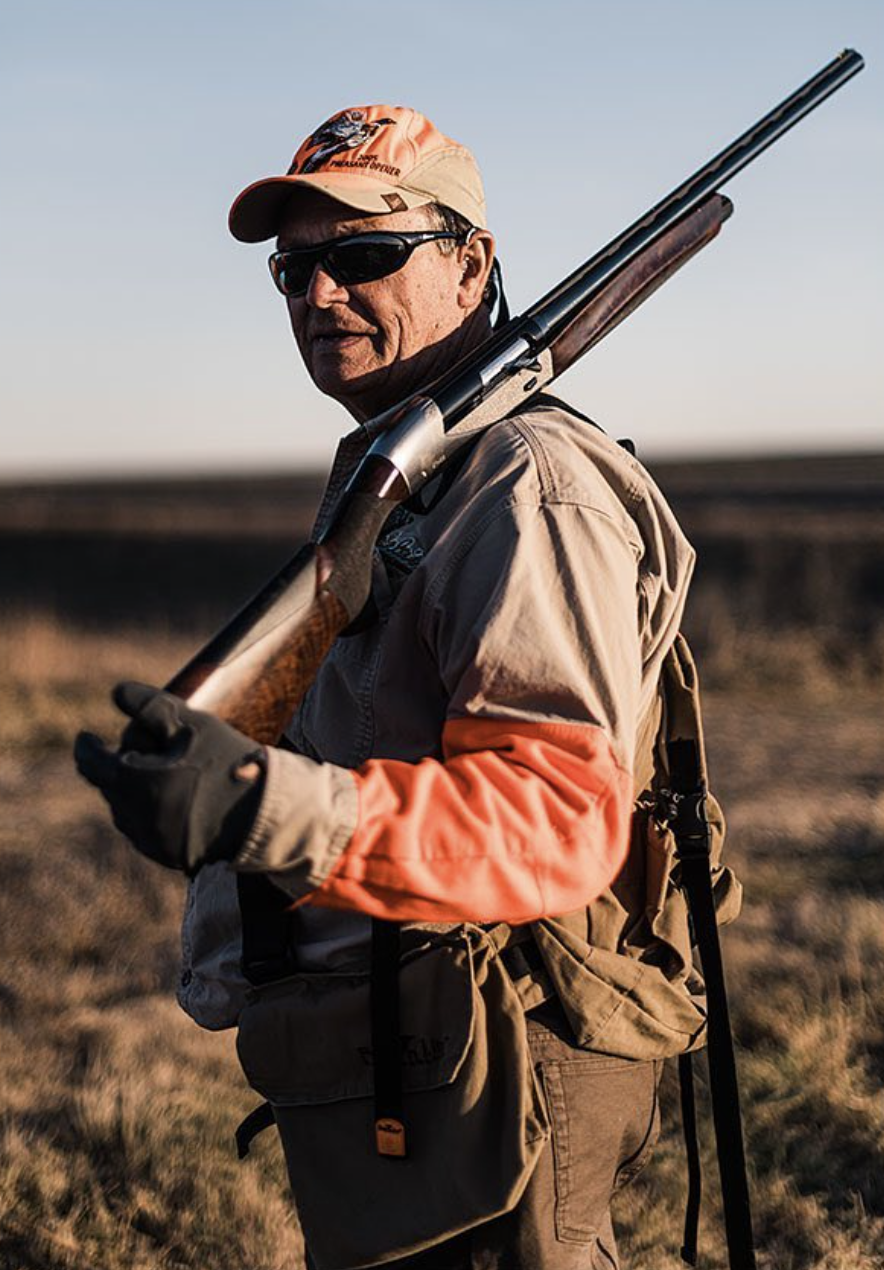 A man wearing sunglasses, a tan cap, and a beige jacket with orange sleeves, holding a shotgun over his shoulder, standing in an open field during sunset.