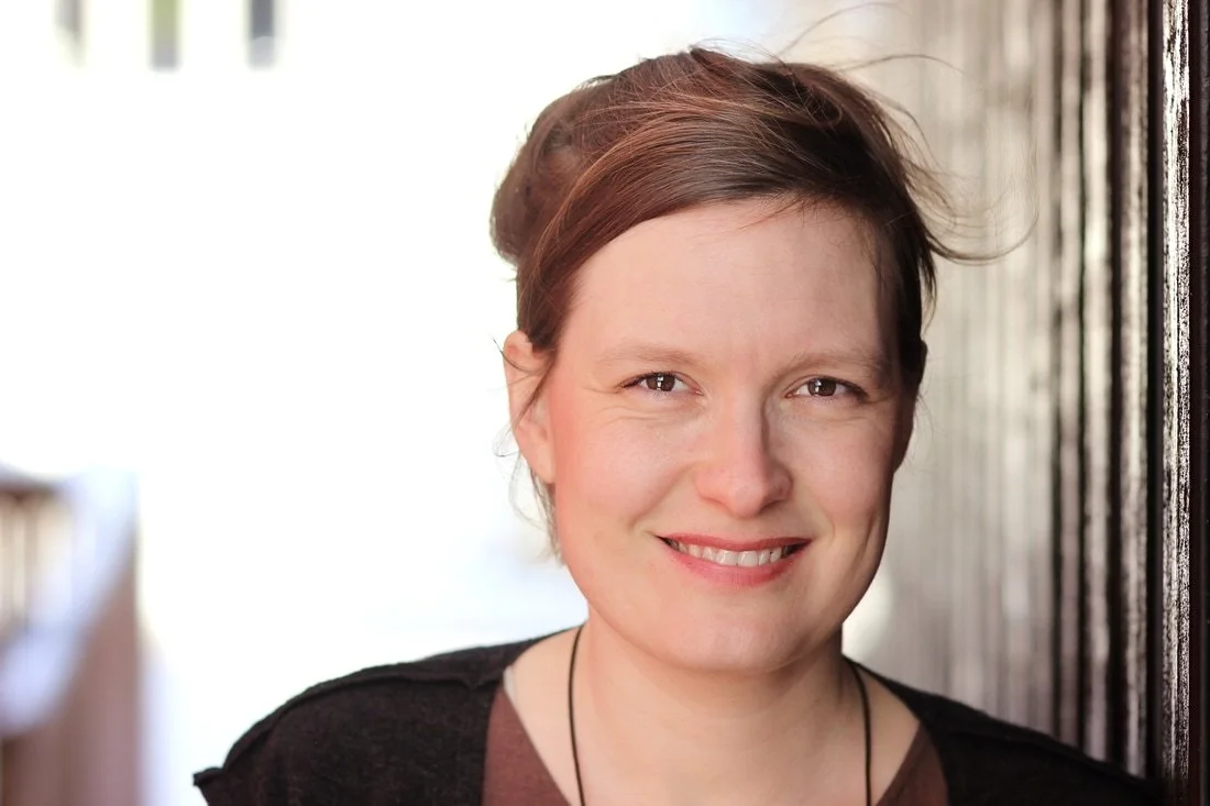 A woman with short brown hair smiling, wearing a black top, standing against a dark textured wall.