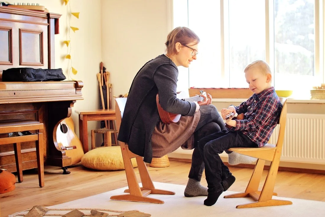 A woman and a young boy sitting inside a bright room by a large window, playing guitars together.