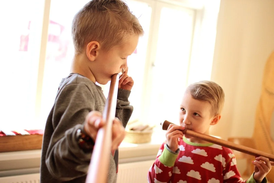Two young boys playing with wooden sticks indoors near a window.