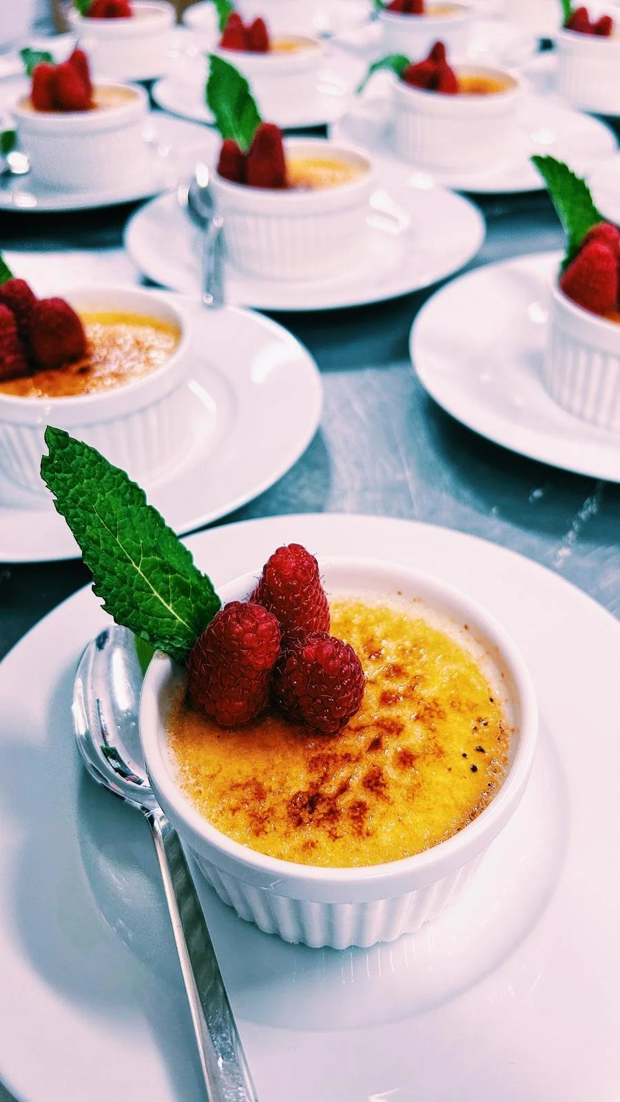 Crème brûlée garnished with raspberries and a mint leaf, served on a white plate with a spoon, multiple similar desserts in the background.
