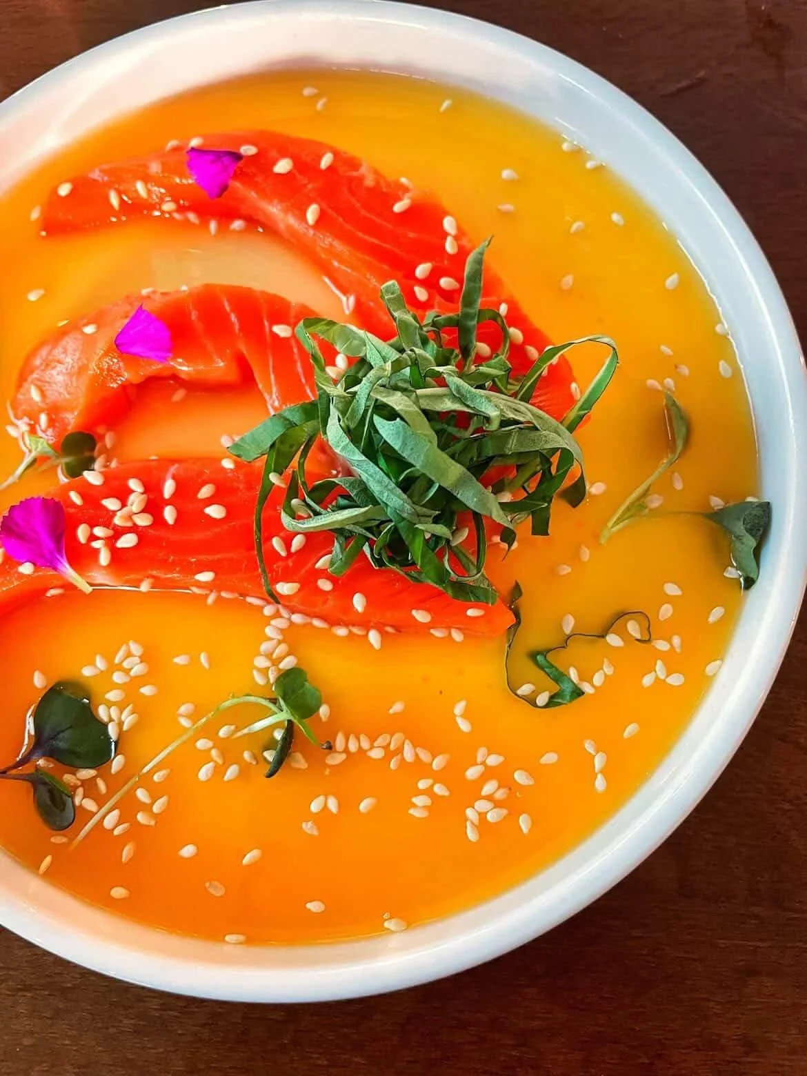 A bowl of salmon sashimi topped with sesame seeds, shredded greens, and purple flower petals, served in a yellow sauce
