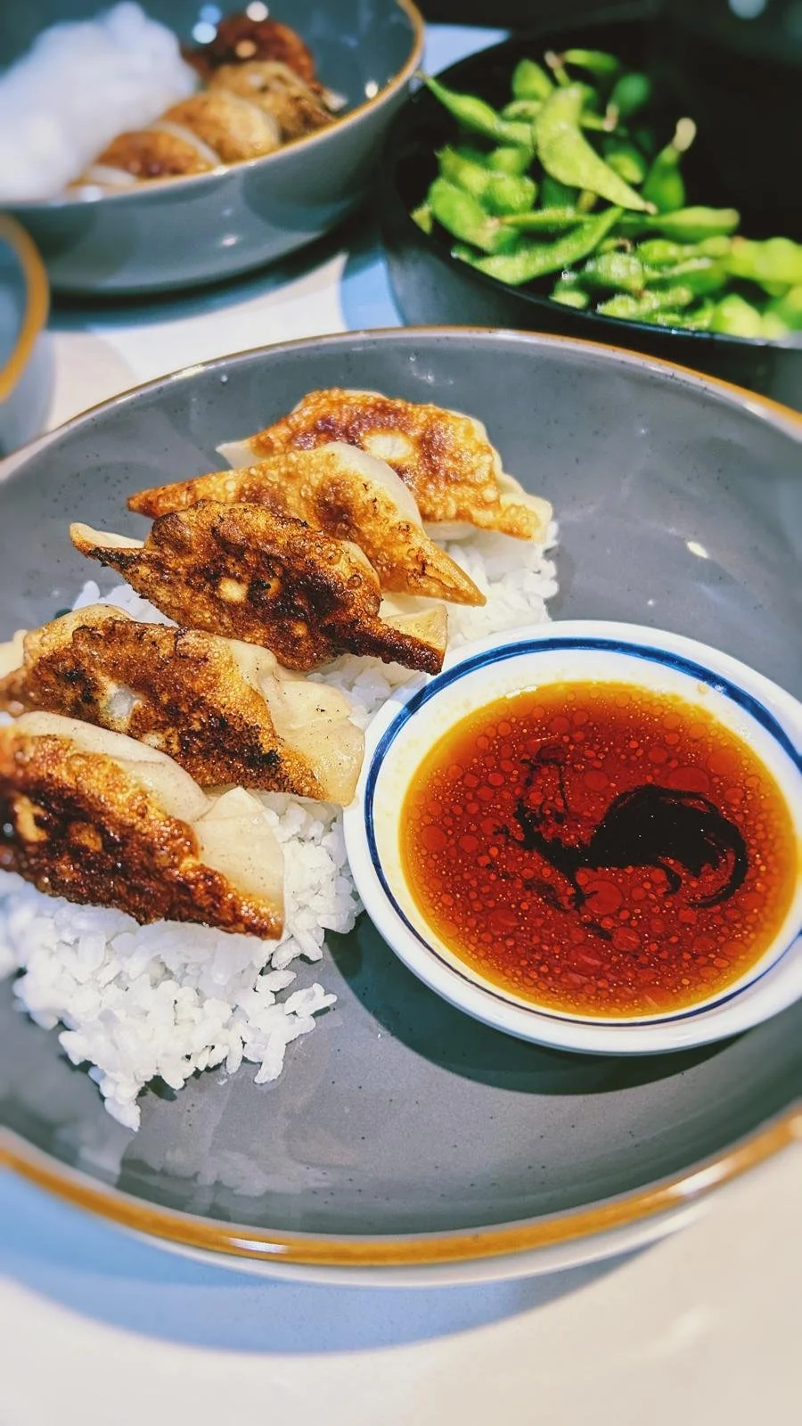 Close-up of a plate with steamed white rice topped with fried dumplings and a small bowl of soy sauce, with bowls of green vegetables and more dumplings in the background.