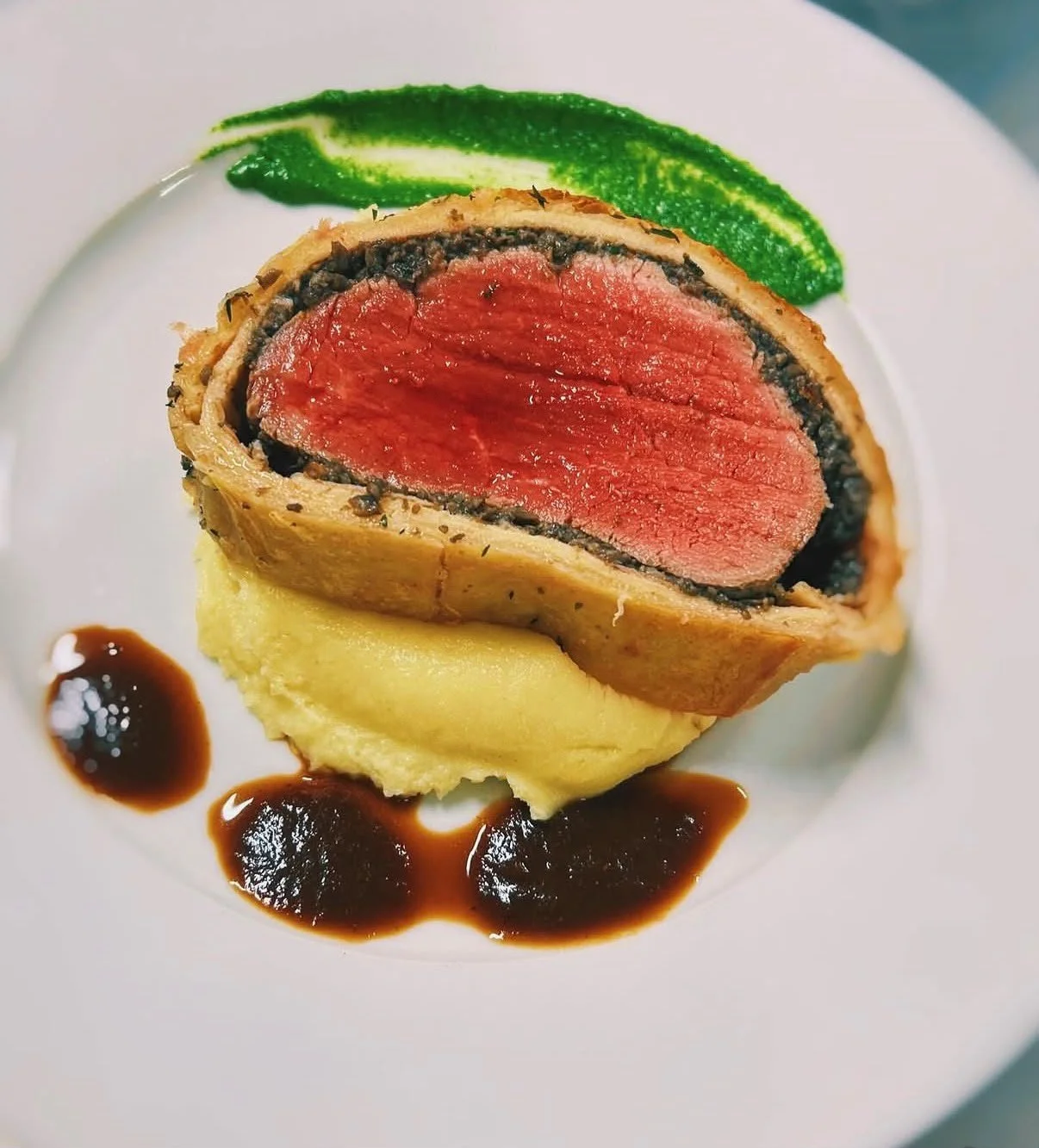 A plated dish featuring a filet mignon wrapped in pastry with a layer of black truffle, served over mashed potatoes, garnished with green sauce and a garlic puree, and drizzled with sauce.