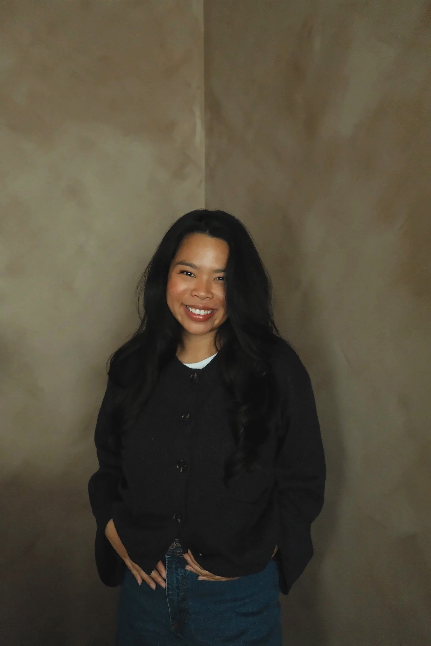 A young woman with long black hair, smiling, wearing a black sweater and blue jeans, standing against a plain beige wall.