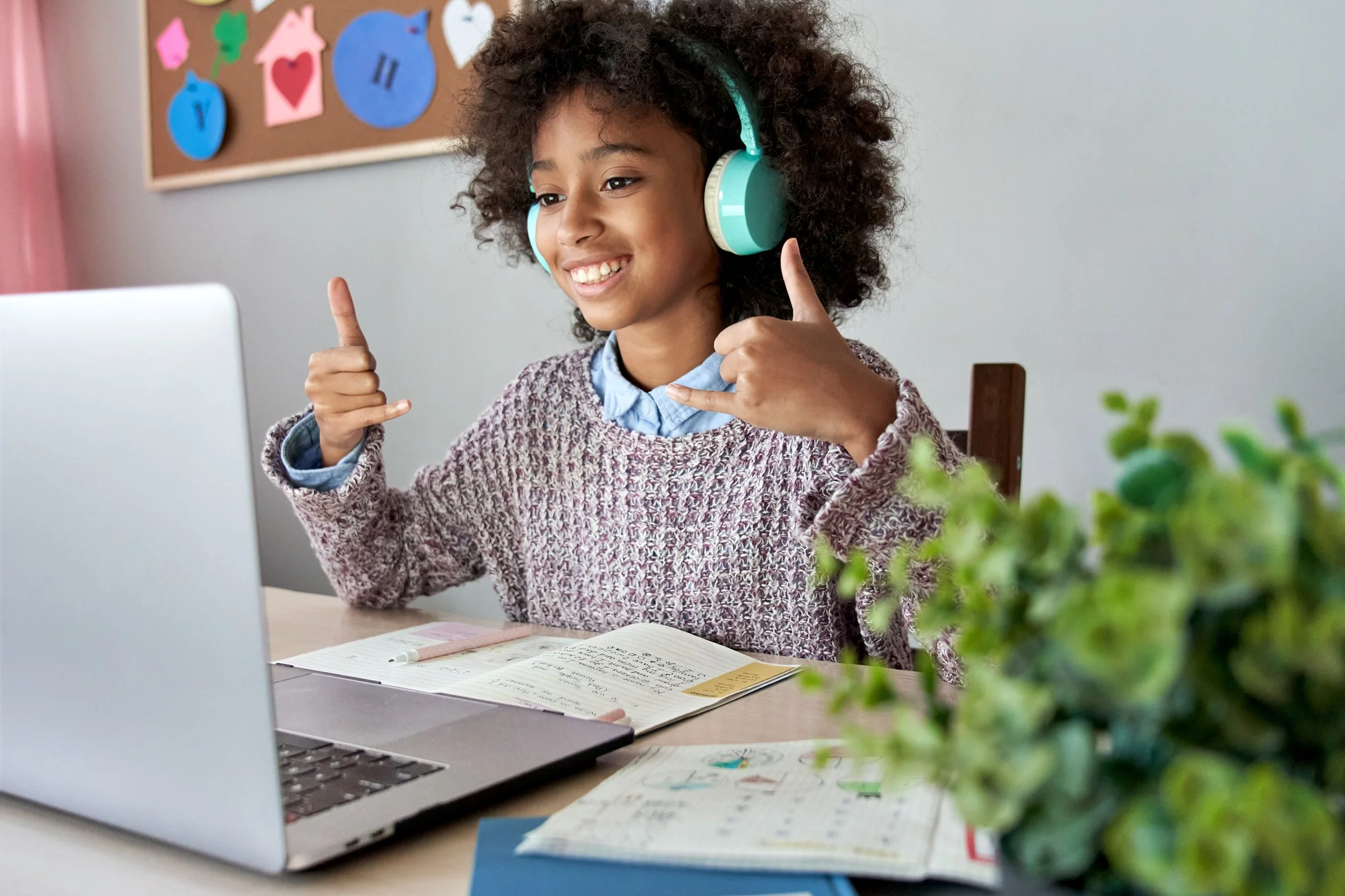 A young girl with curly hair wearing headphones, smiling and participating in a speech therapy teletherapy visit. There is a bulletin board with colorful paper cutouts in the background and a potted plant in the foreground