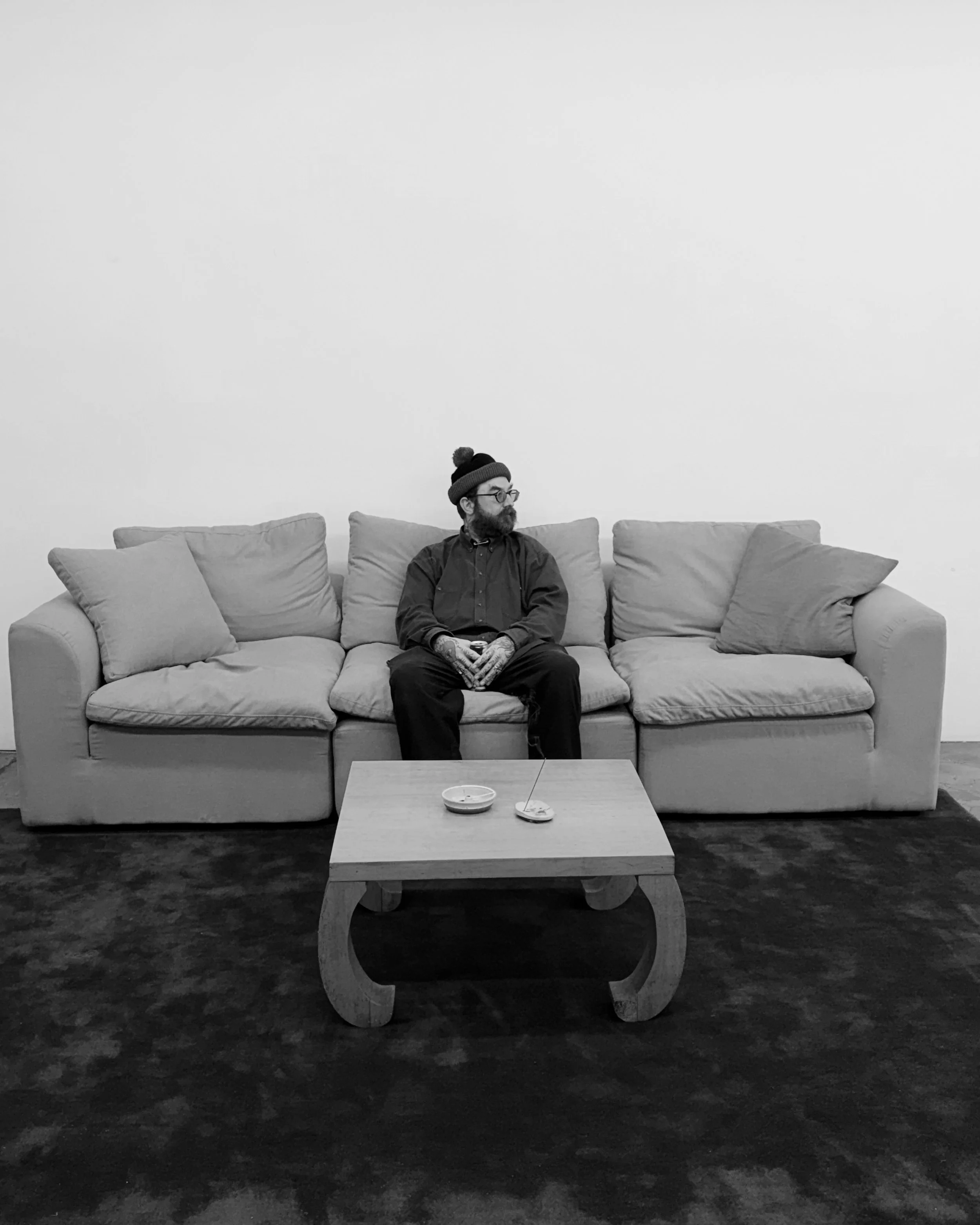 Bearded man wearing glasses and a knit beanie sitting on a couch behind a coffee table
