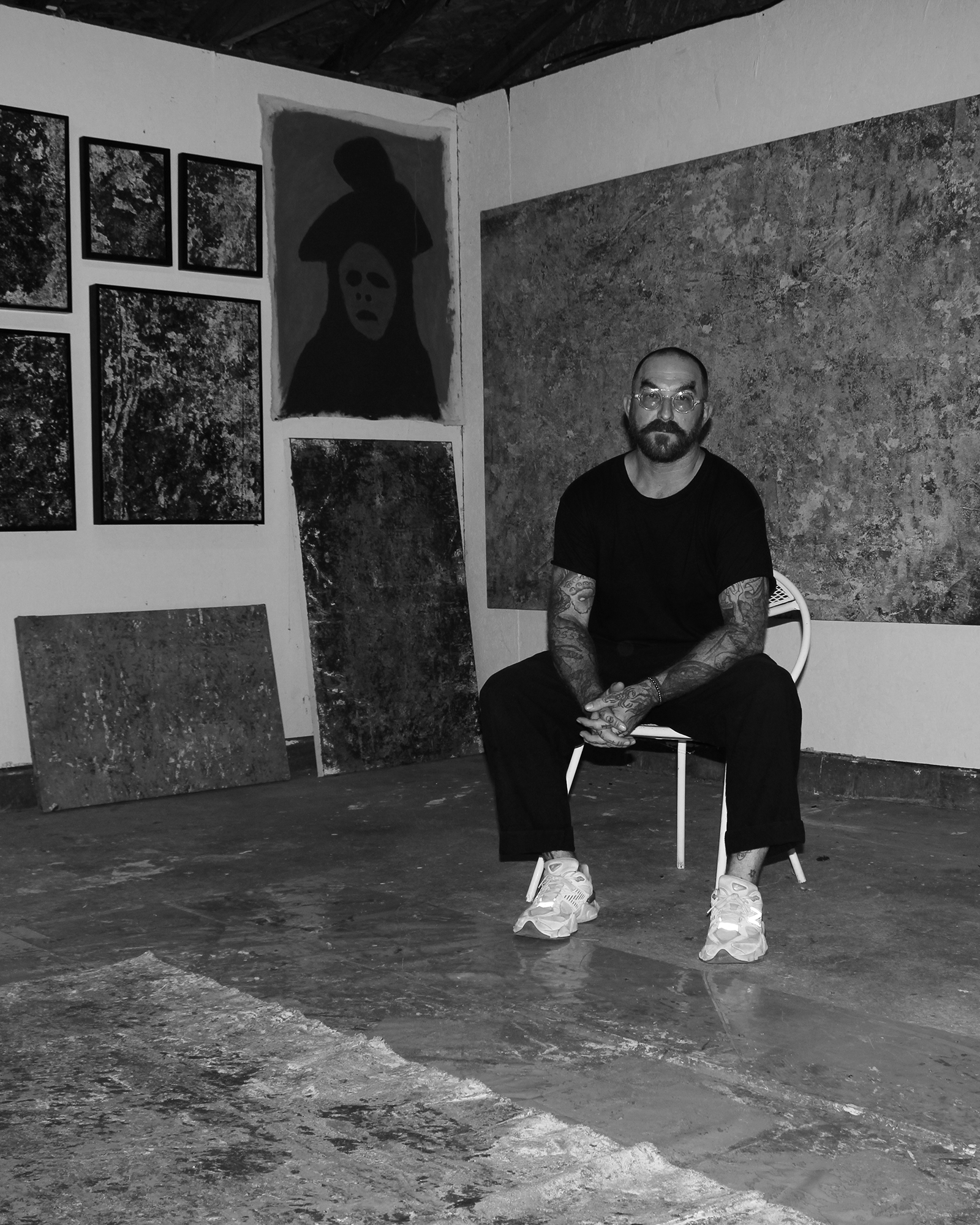 Bearded tattooed man sitting in an art studio surrounded by abstract paintings and a portrait.