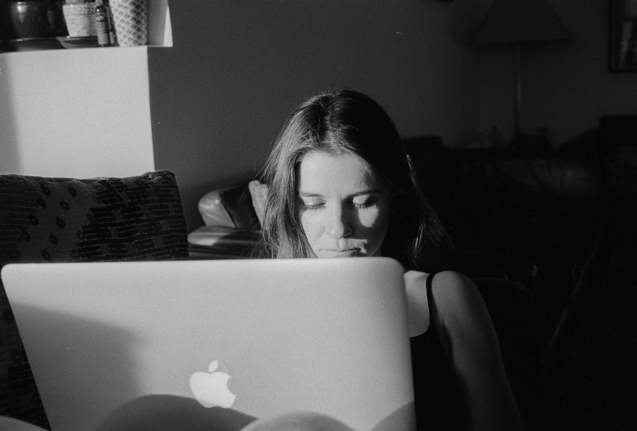 A black and white photo of a woman with her eyes closed, sitting in front of an open laptop, in a dimly lit room with furniture and objects in the background.