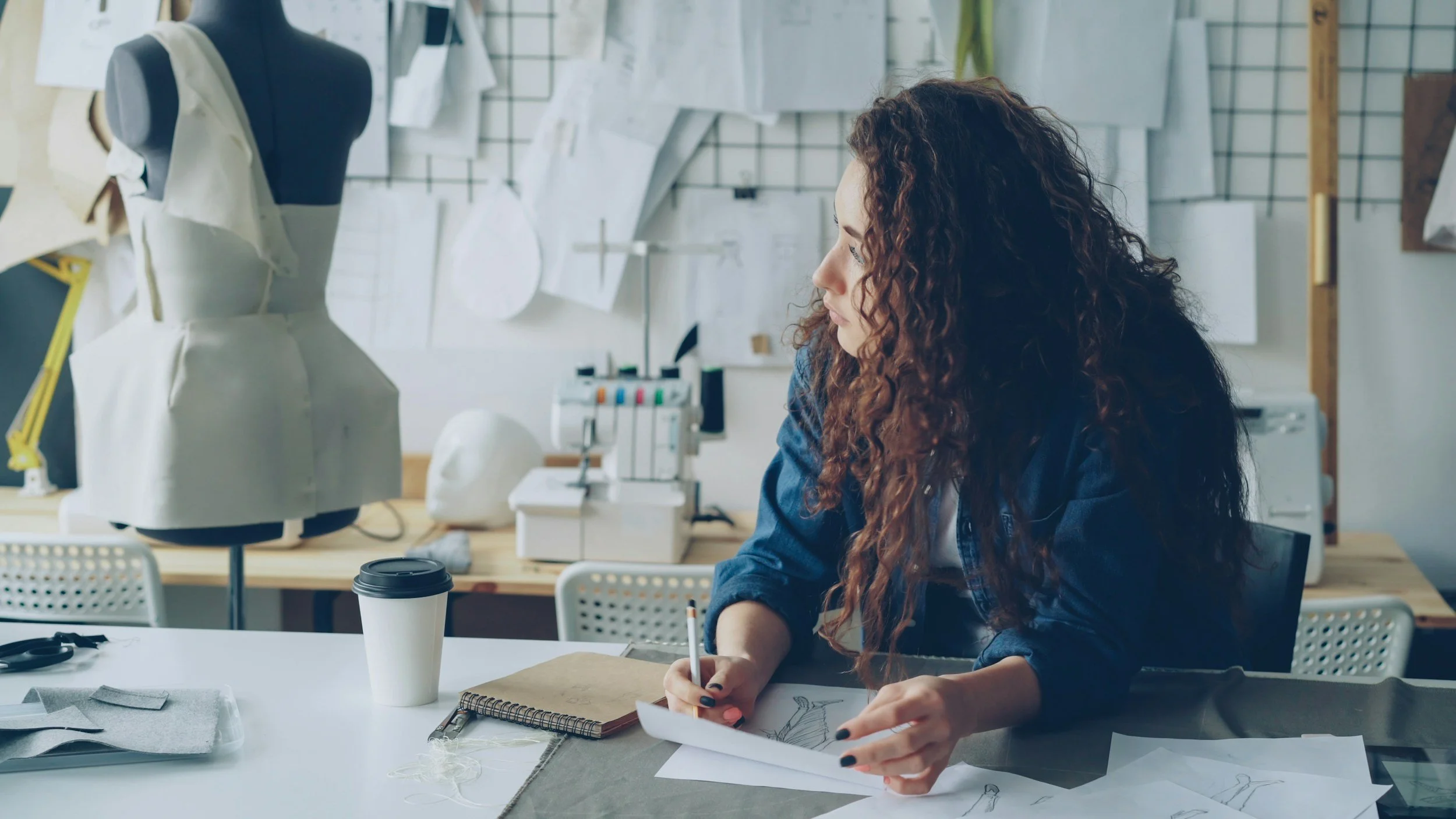 A woman with long curly hair sitting at a table in an art studio, sketching designs on paper, with sewing materials and a mannequin in the background.