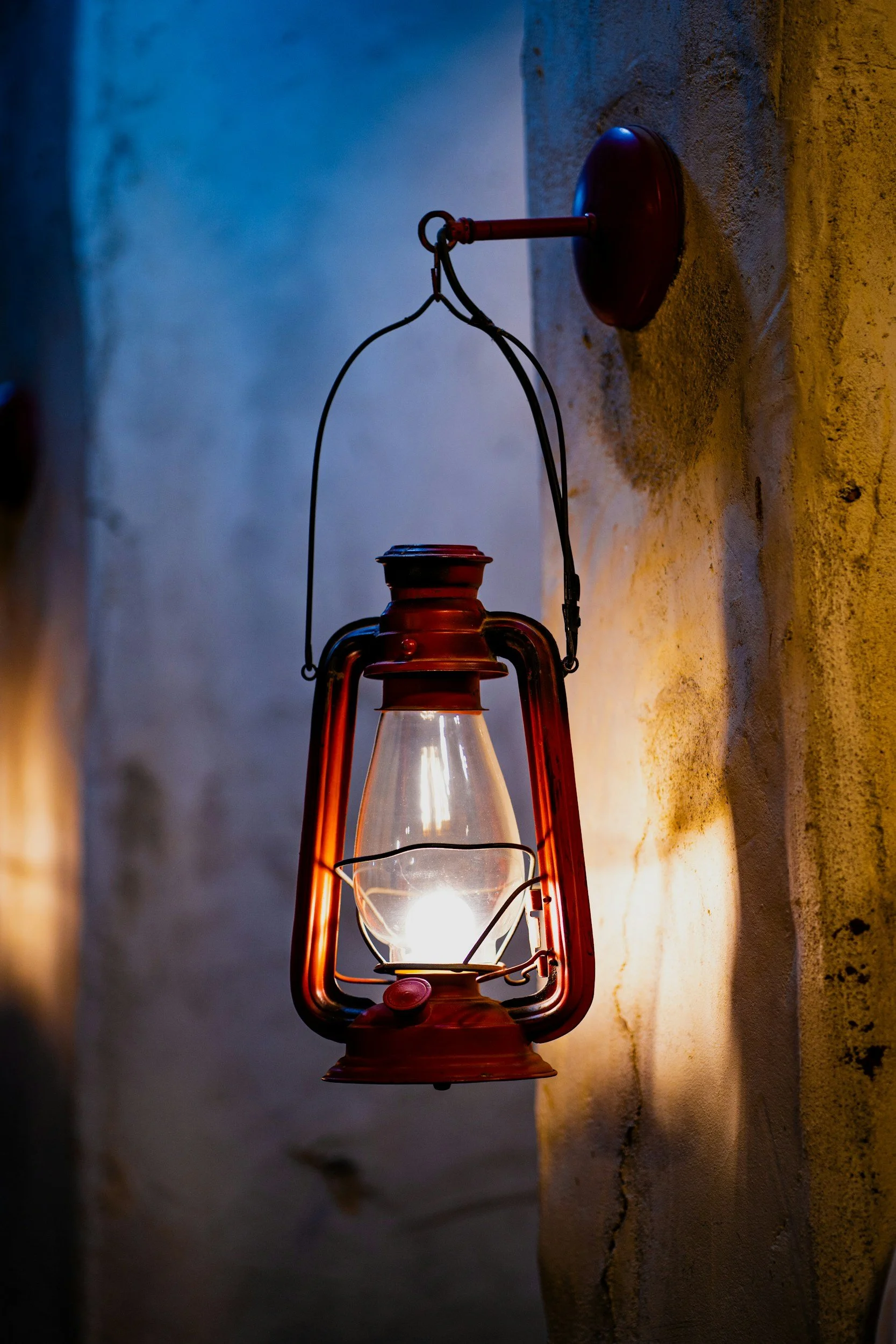 A vintage red oil lantern hanging on a textured stucco wall, illuminated and casting a warm glow.