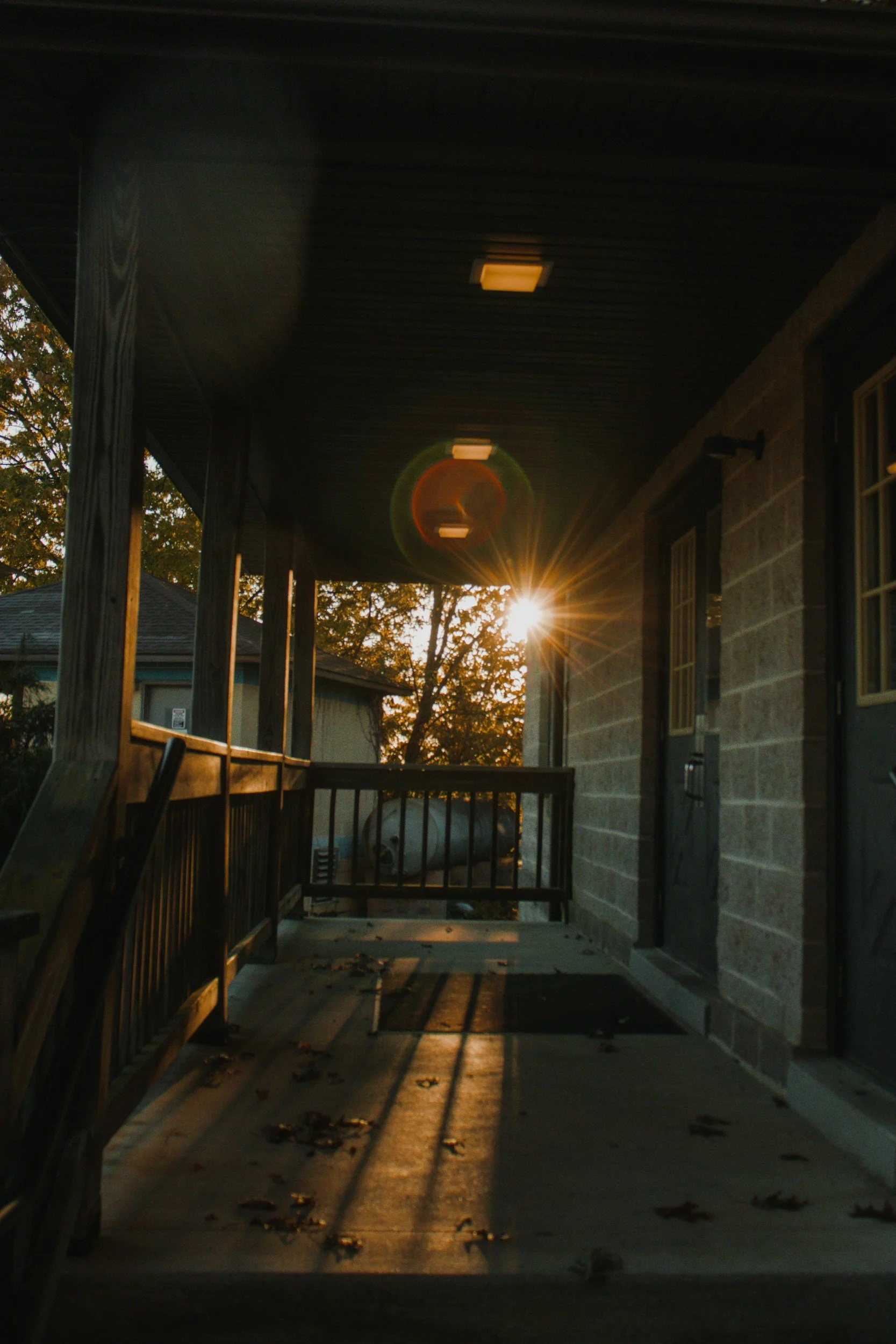 Sunset viewed from a house porch with a wooden railing, overhead lights, and falling leaves on the concrete floor.