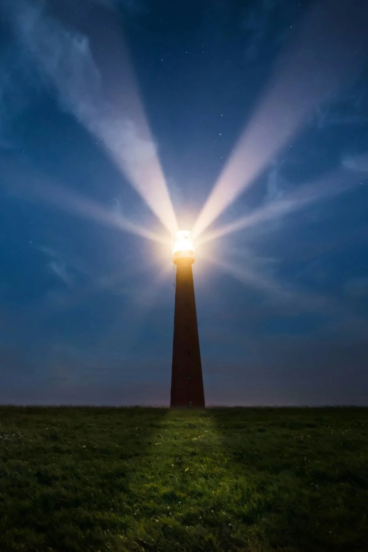 Nighttime photo of a lighthouse with its light shining, creating beams that extend outward into the sky, surrounded by a dark blue sky with some stars visible.