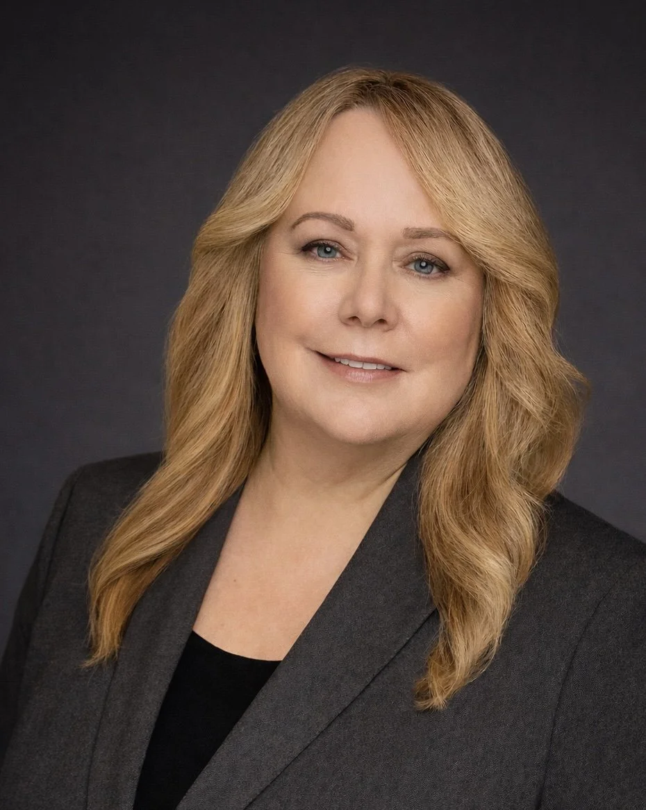 Professional woman with blonde wavy hair and light blue eyes wearing a dark blazer and black top against a dark background.