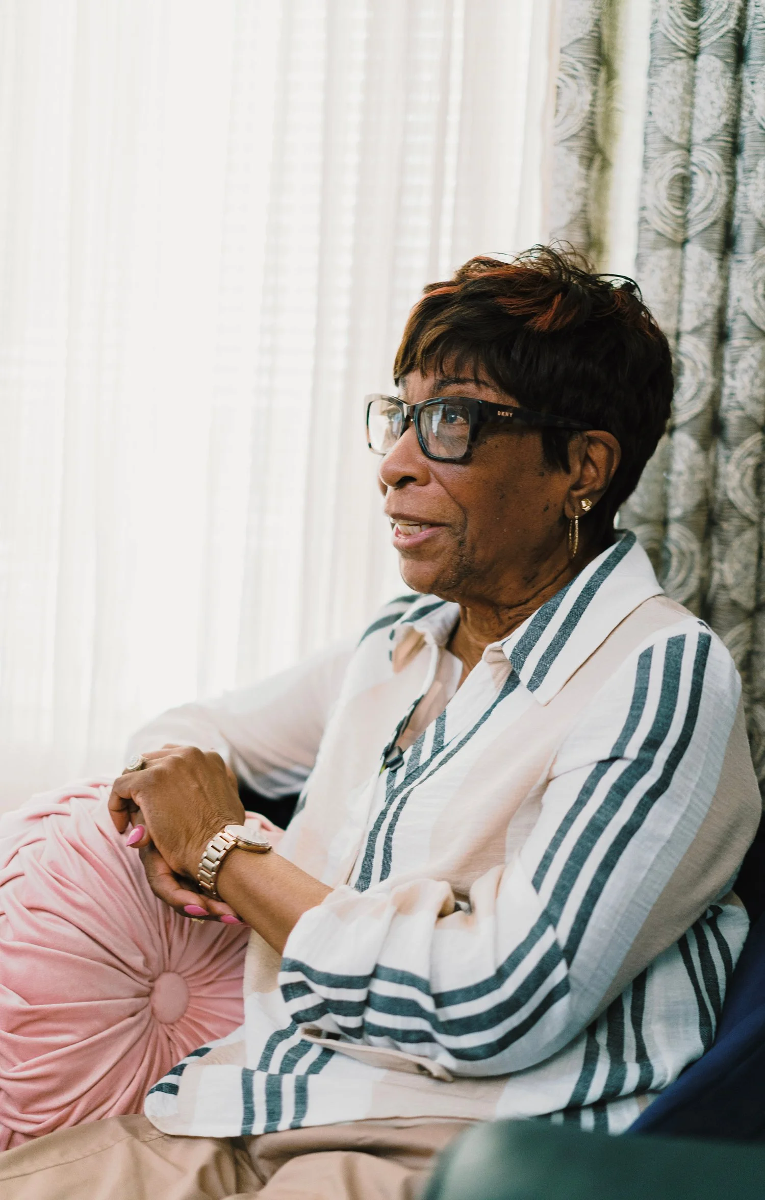 An elderly woman with short dark hair wearing glasses and a striped shirt, smiling and sitting indoors near curtains.