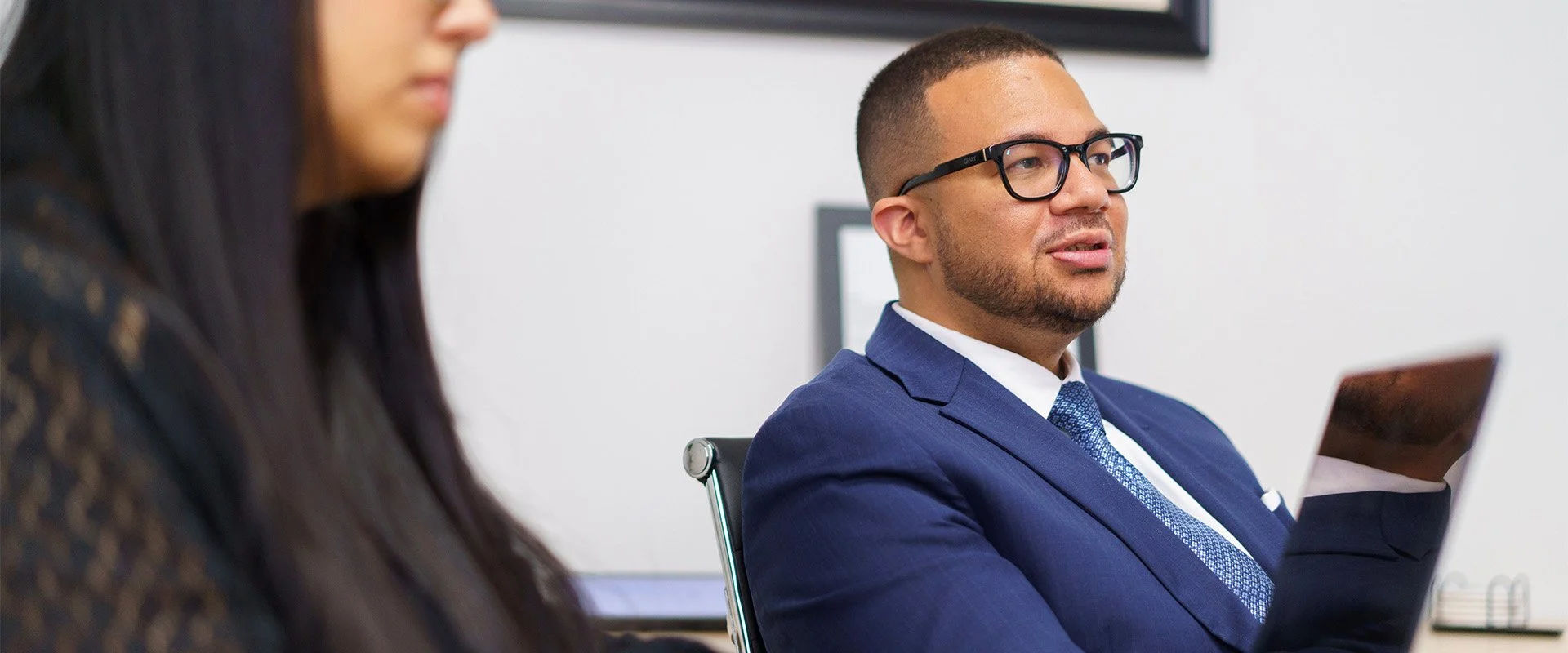 Raphael in a blue suit and glasses sitting in an office, holding a tablet, with a woman in the foreground partially visible.