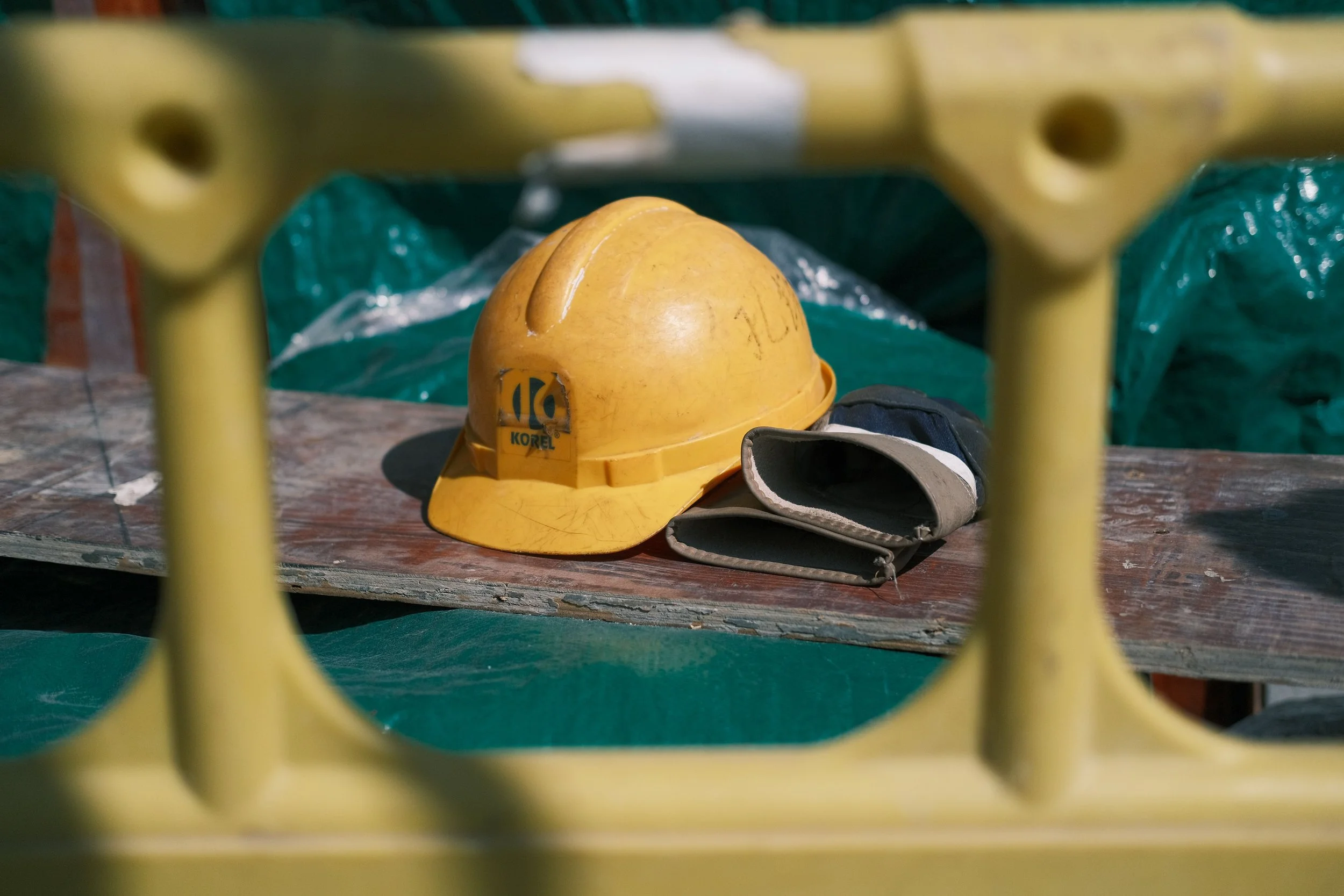 A yellow safety helmet and safety goggles are placed on a wooden surface, seen through yellow safety barrier rails at a construction site.