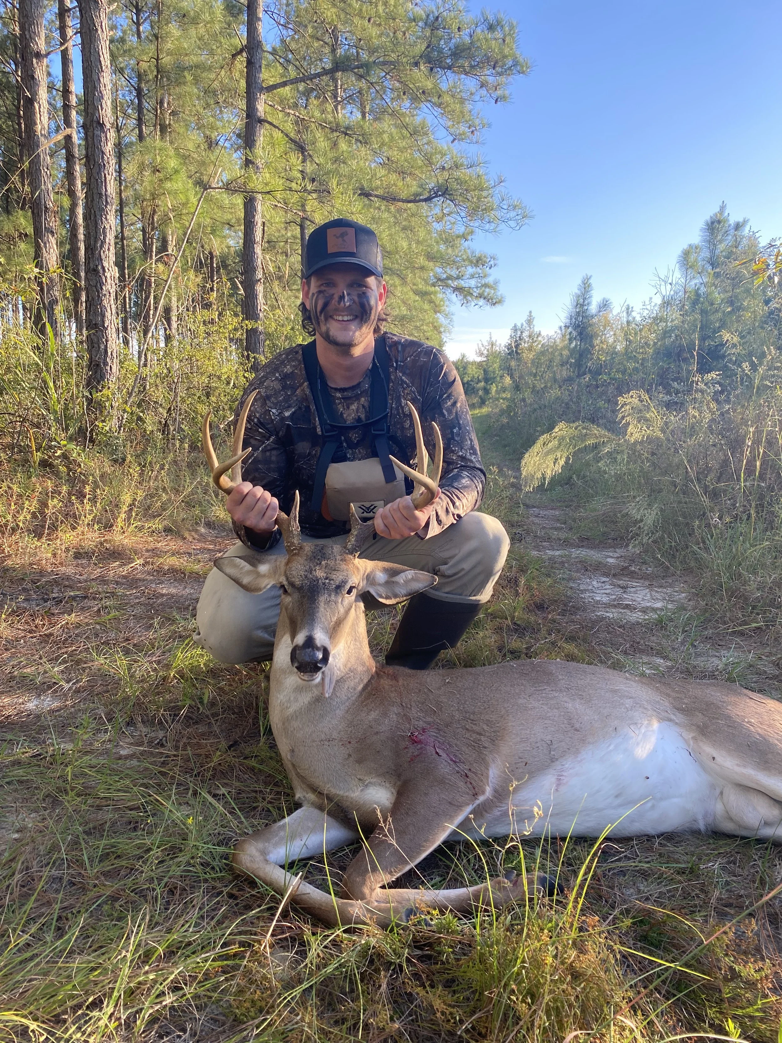 Hunter in camouflage holding a harvested antlered deer in a pine forest
