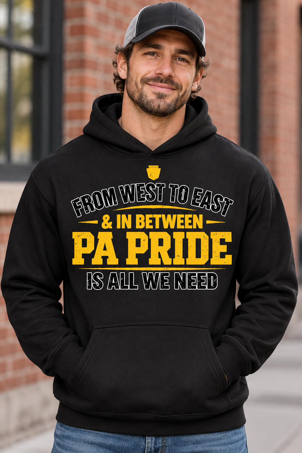 A man wearing a black hoodie with yellow and white text that reads 'From West to East & in Between PA Pride is All We Need,' standing outdoors in front of a brick building.